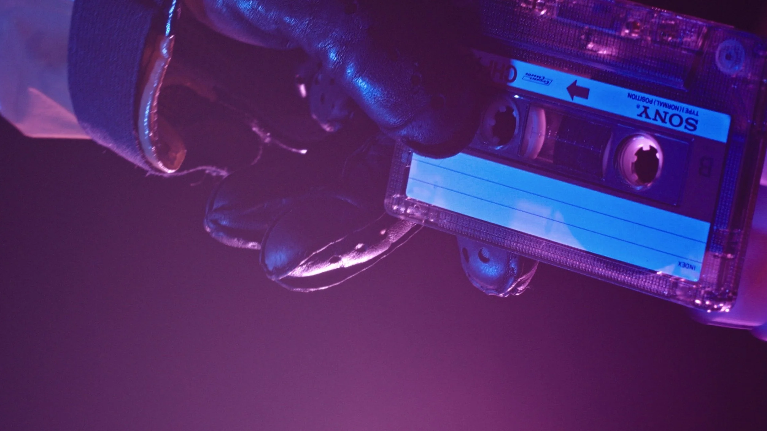 Gloved hands holding a Sony cassette tape under purple lighting