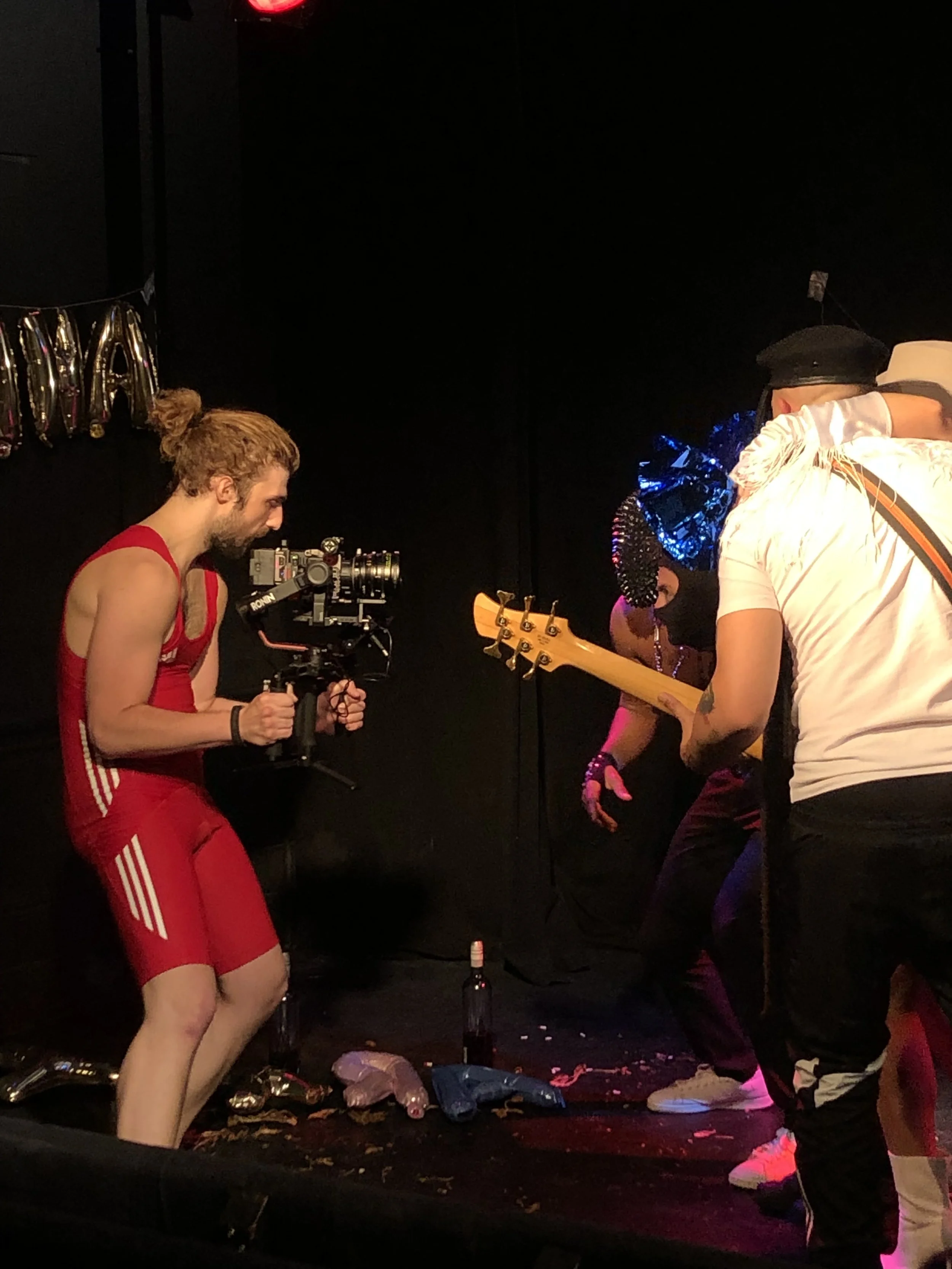 A person in a red outfit films a group on stage playing instruments. They hold a camera, while the group is adorned in colorful costumes under stage lighting.