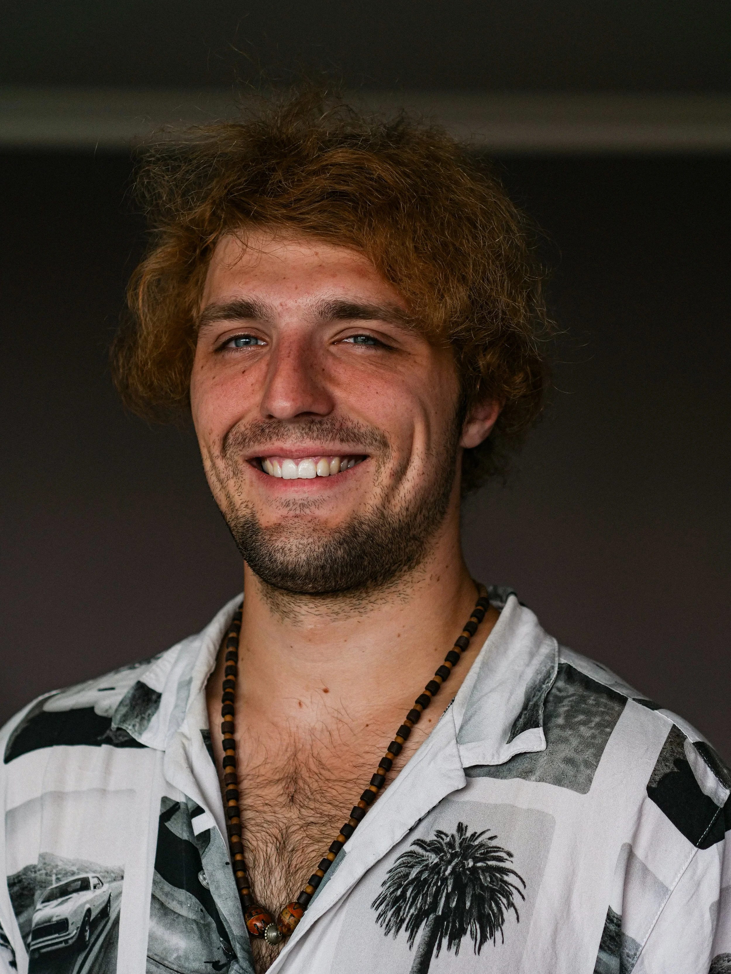 Smiling man with curly hair wearing a patterned shirt and beaded necklace