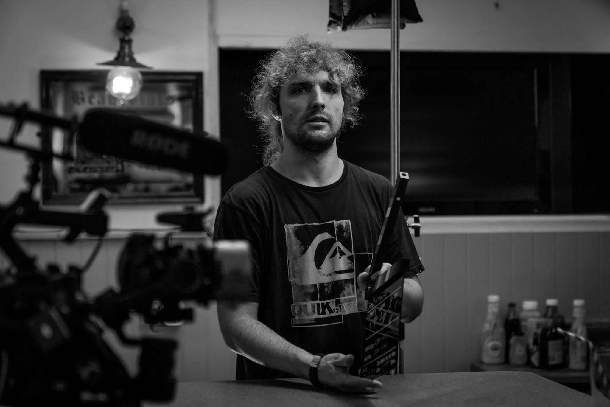 Black and white photo of a person holding a clapperboard on a film set, with a microphone and camera visible in the foreground. Condiment bottles are in the background.