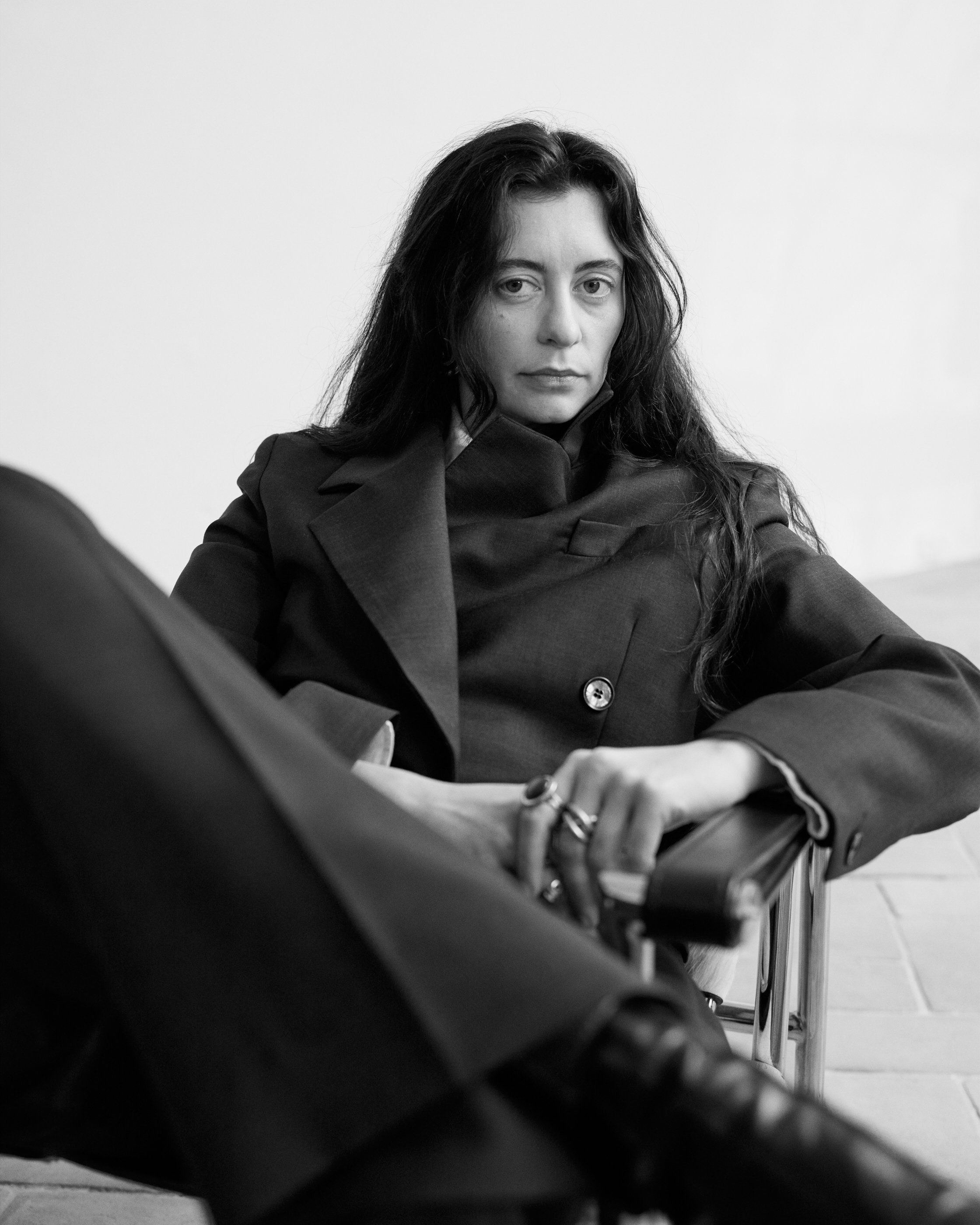 A woman with long curly hair sitting on a chair, wearing a dark coat, looking directly at the camera with a serious expression.