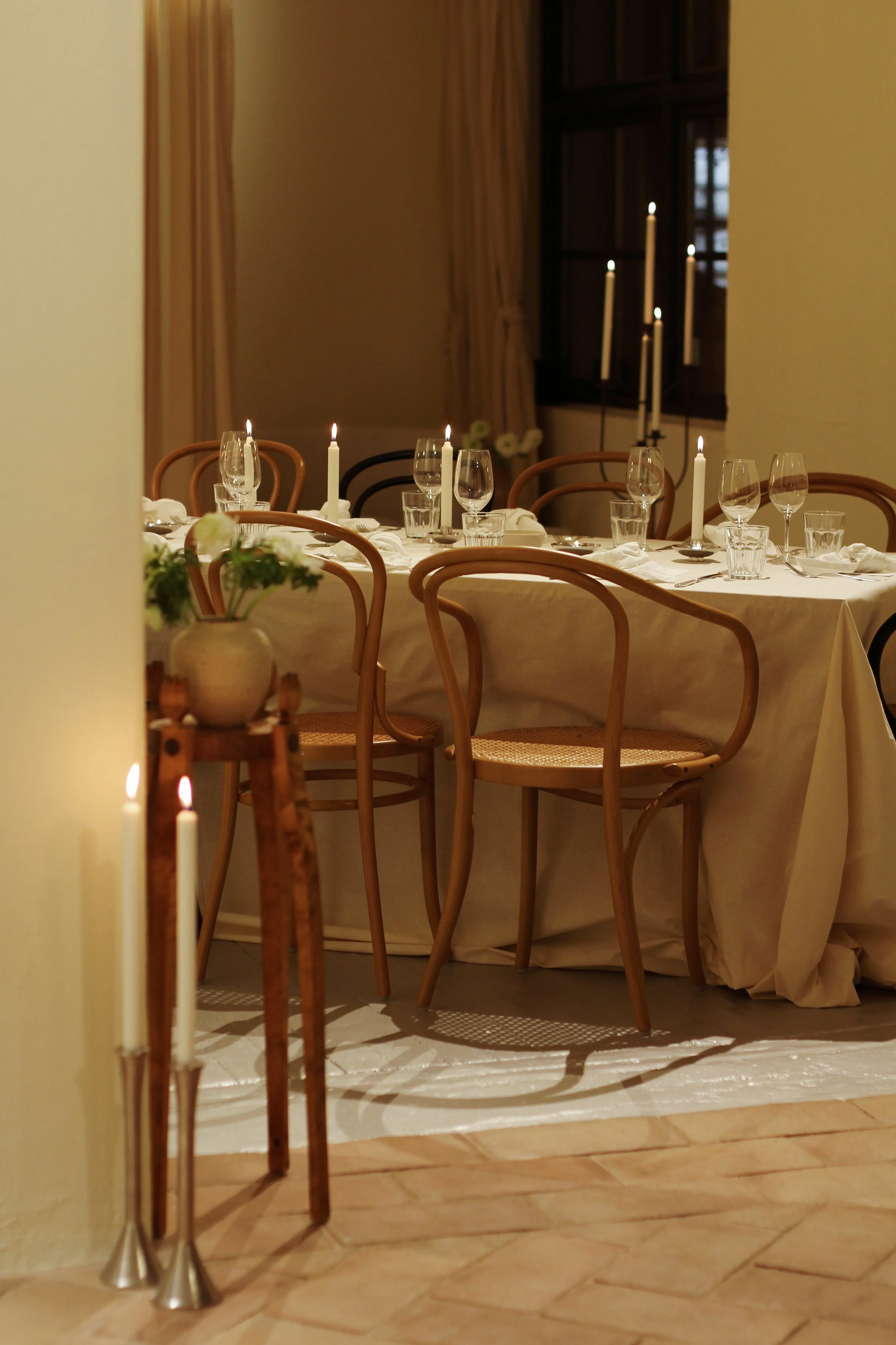 A dinner table set with wine glasses, water glasses, candles, and napkins in a warmly lit room, with chairs around the table and a window in the background.