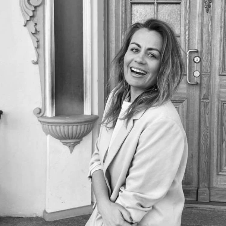 A woman with wavy hair smiling and looking at the camera, standing in front of a wooden door and decorative wall shelf in black and white.
