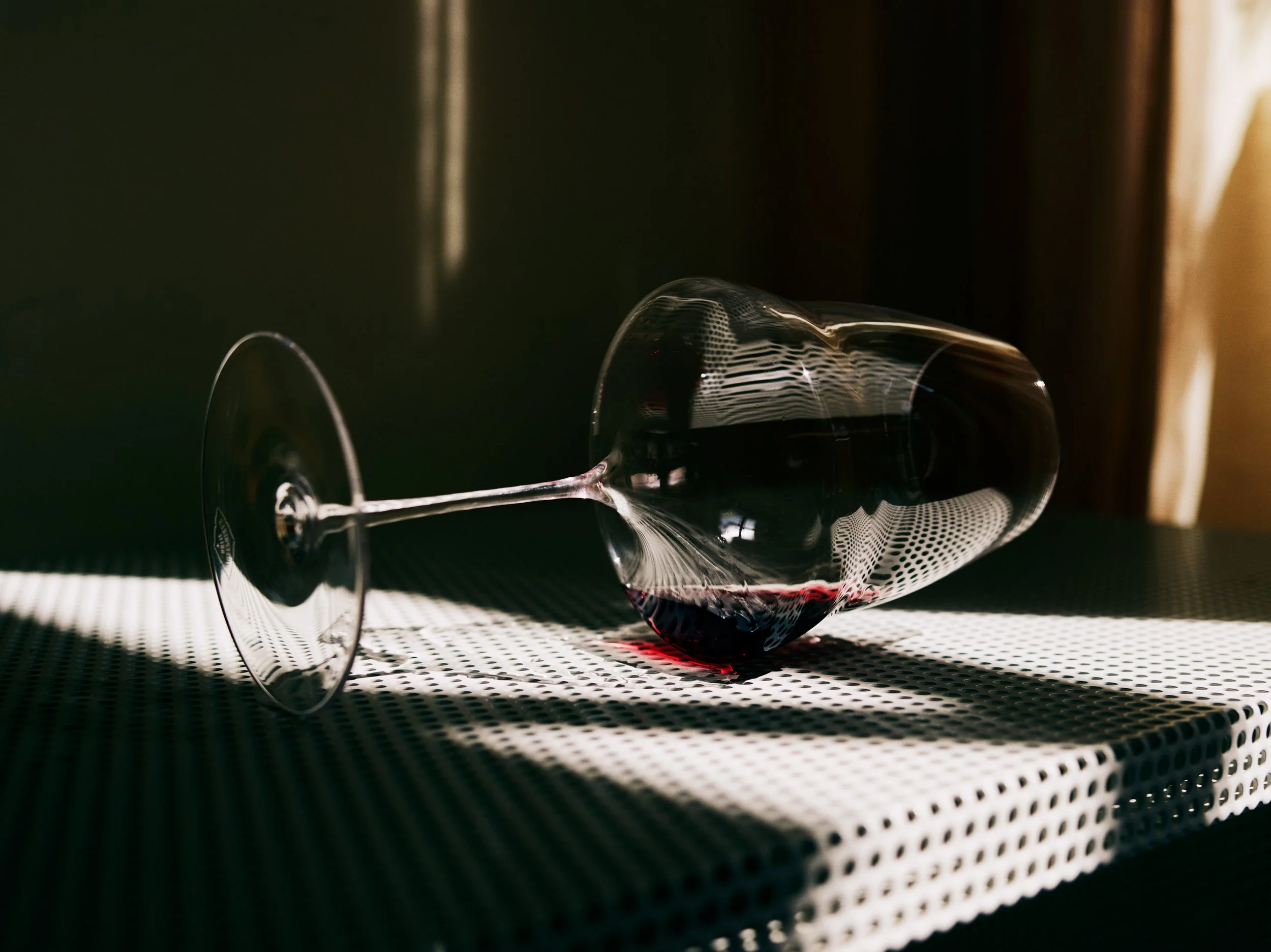 An overturned wine glass with red wine spilled on a textured surface, with shadows and light creating a dramatic effect.