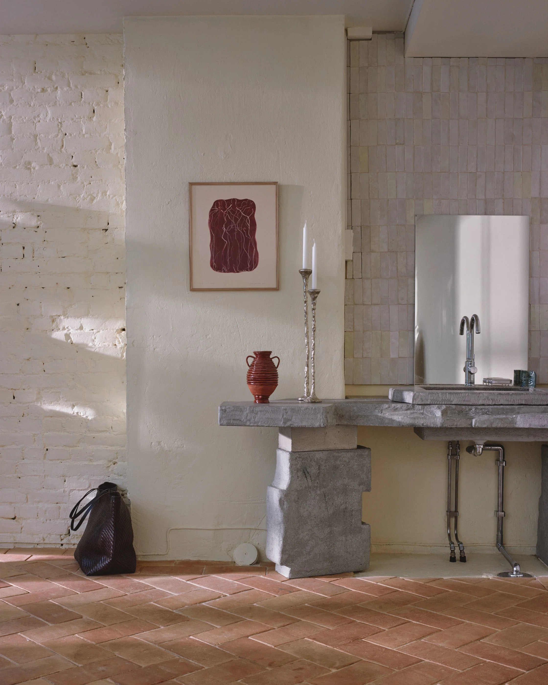 Interior of a room featuring a white brick wall, artwork with a red background, and a concrete countertop with a faucet, candlesticks, and a small vase. A brown bag rests on the floor near the wall.