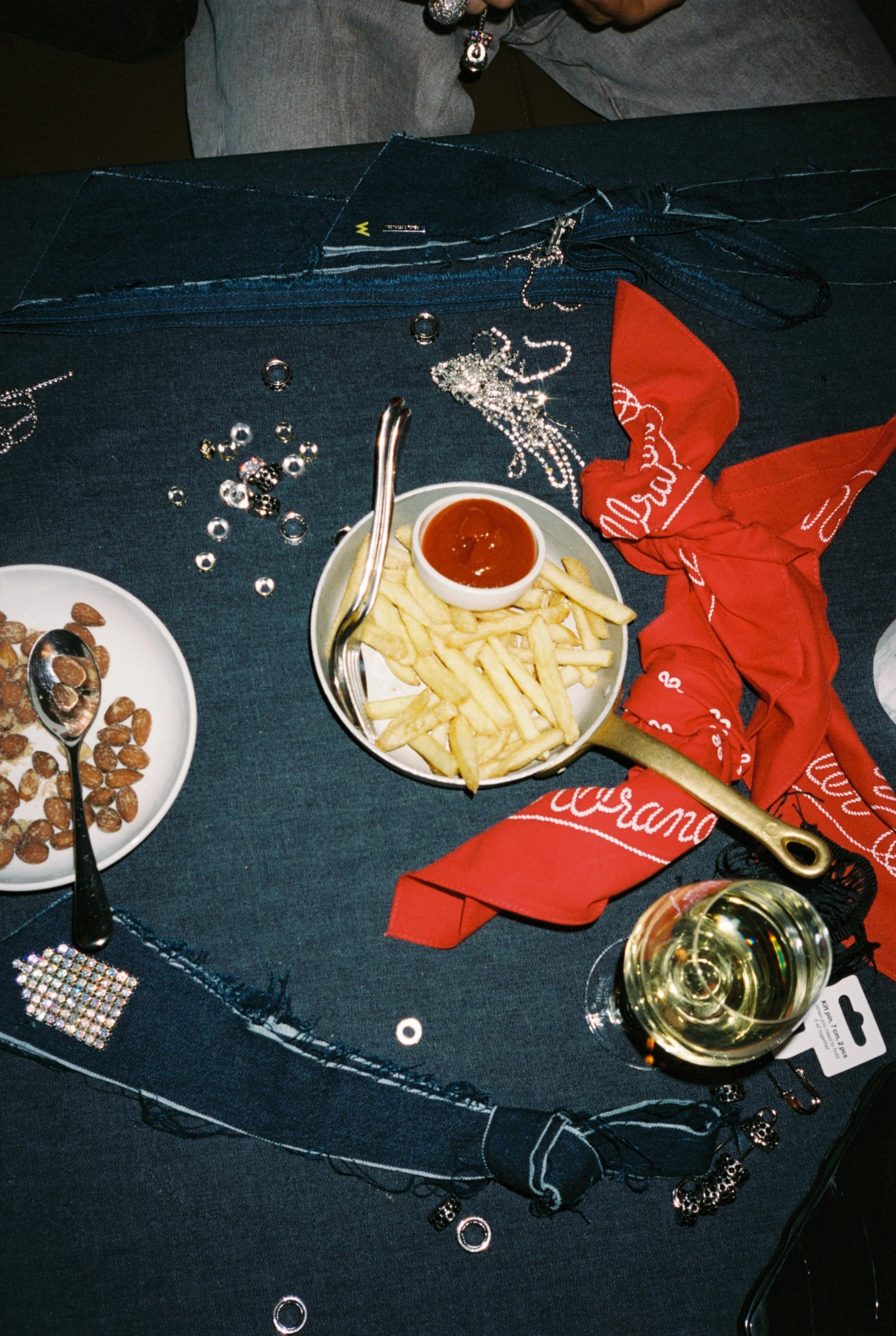 A table with a small plate of French fries with ketchup, a bowl of mixed nuts, and a glass of white wine, surrounded by jewelry, a red bandana, and scattered sequins.