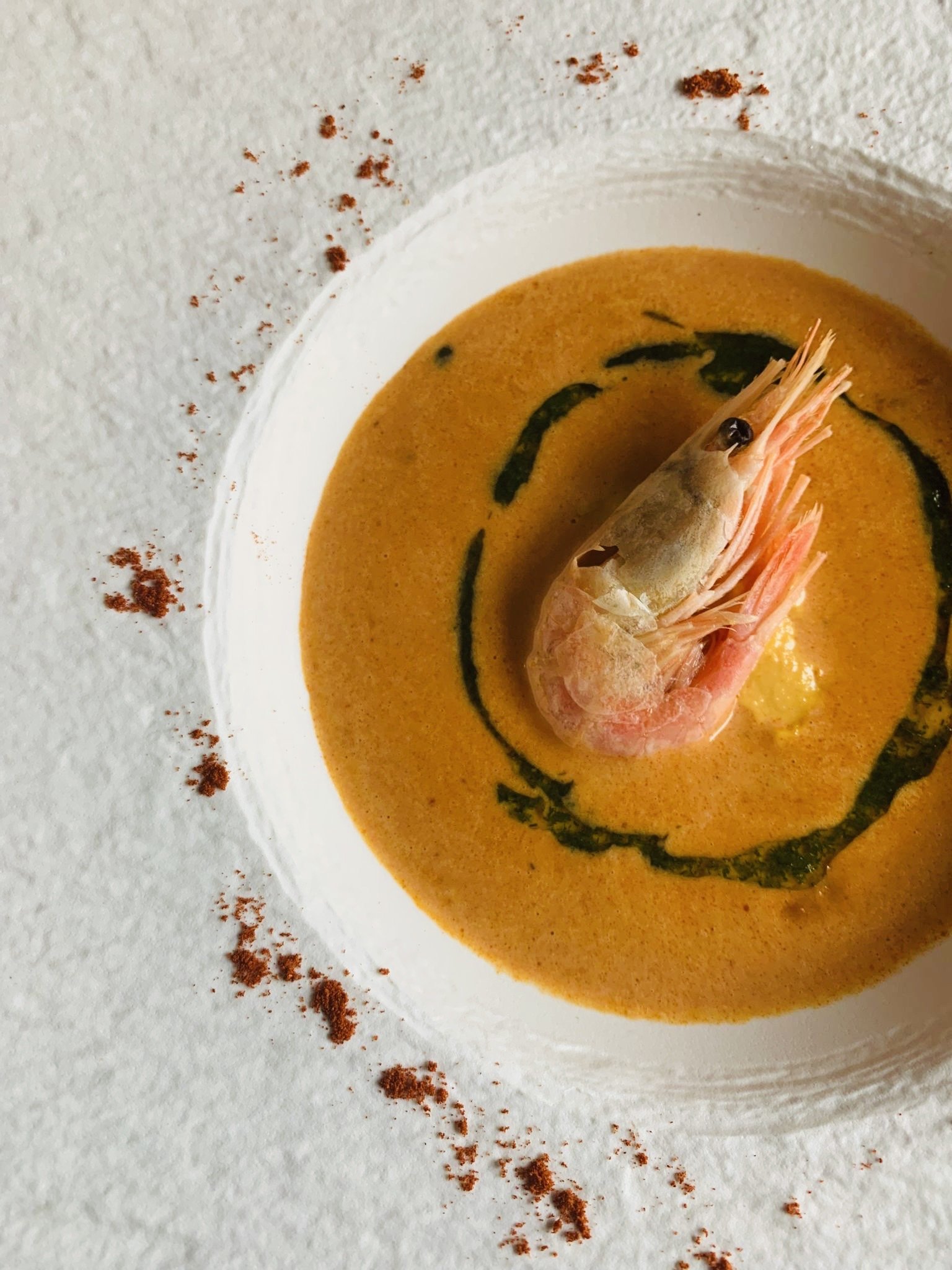 A bowl of creamy orange seafood soup garnished with a cooked shrimp and drizzled with green oil, presented on a textured white surface with red spice sprinkled around the bowl.