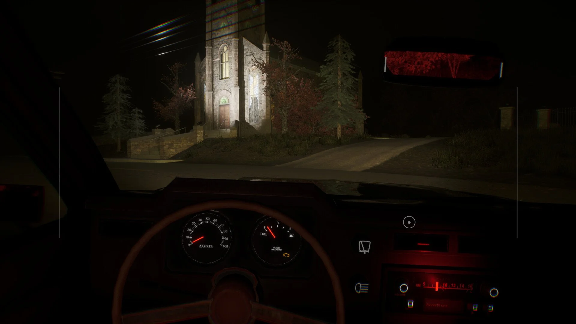 Nighttime view from inside a car showing a church with stone walls, a wooden door, and tall windows, surrounded by trees and a road leading to the church.
