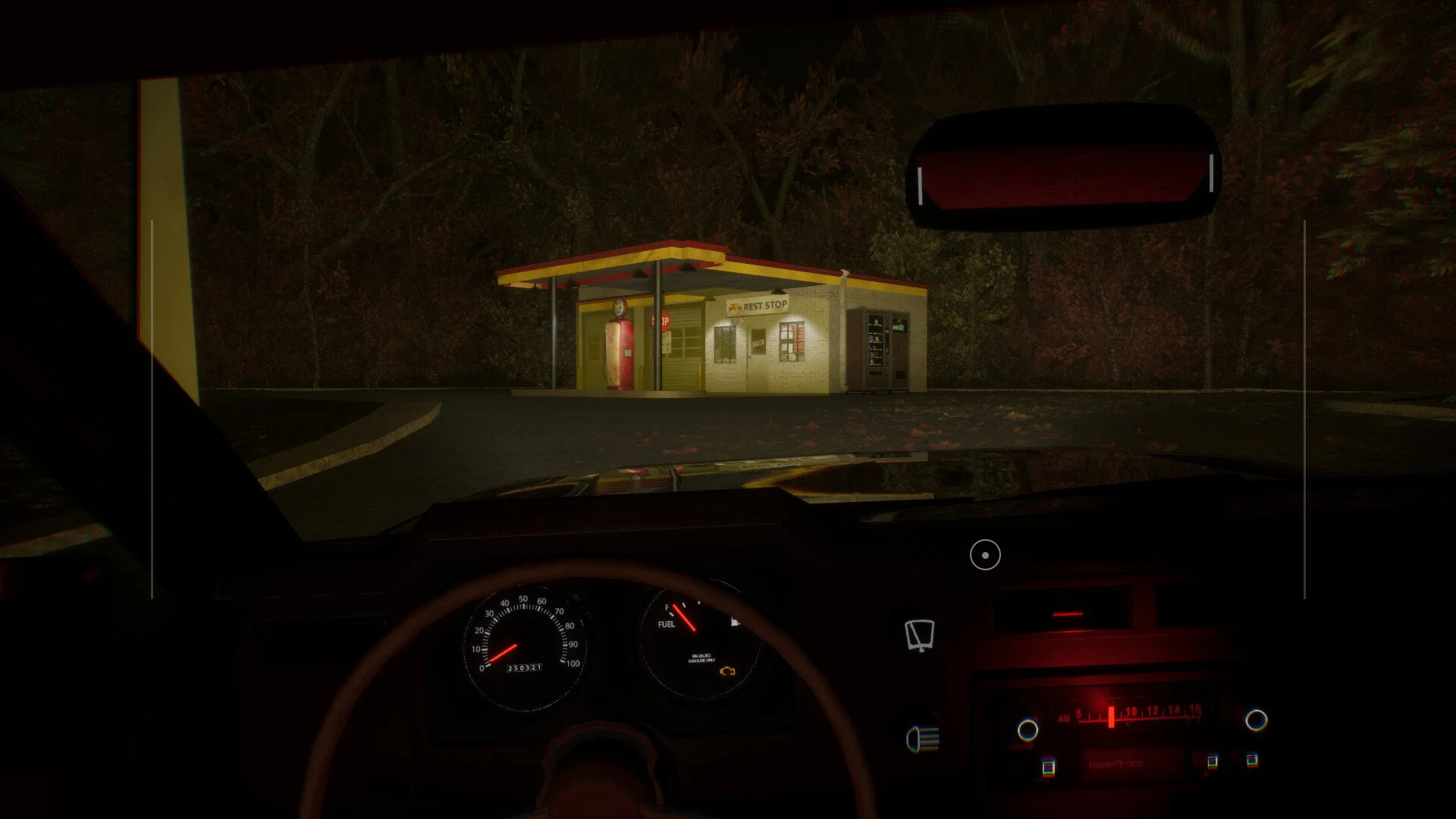 View from inside a car at night looking towards a small yellow and red rest stop building with a gas pump and vending machines, surrounded by trees.