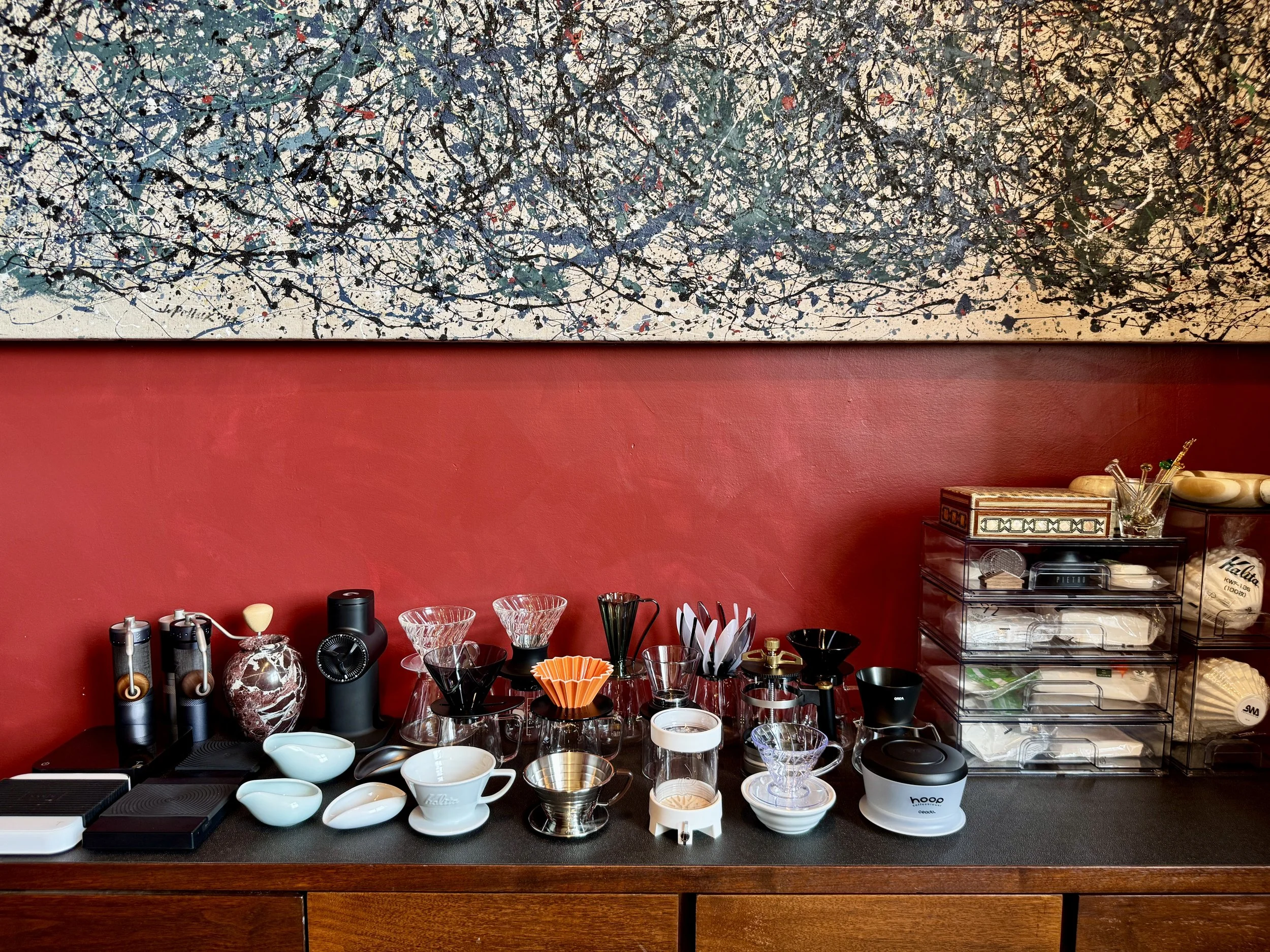 Coffee brewing accessories and cups on a black countertop against a red wall with a colorful abstract painting overhead.