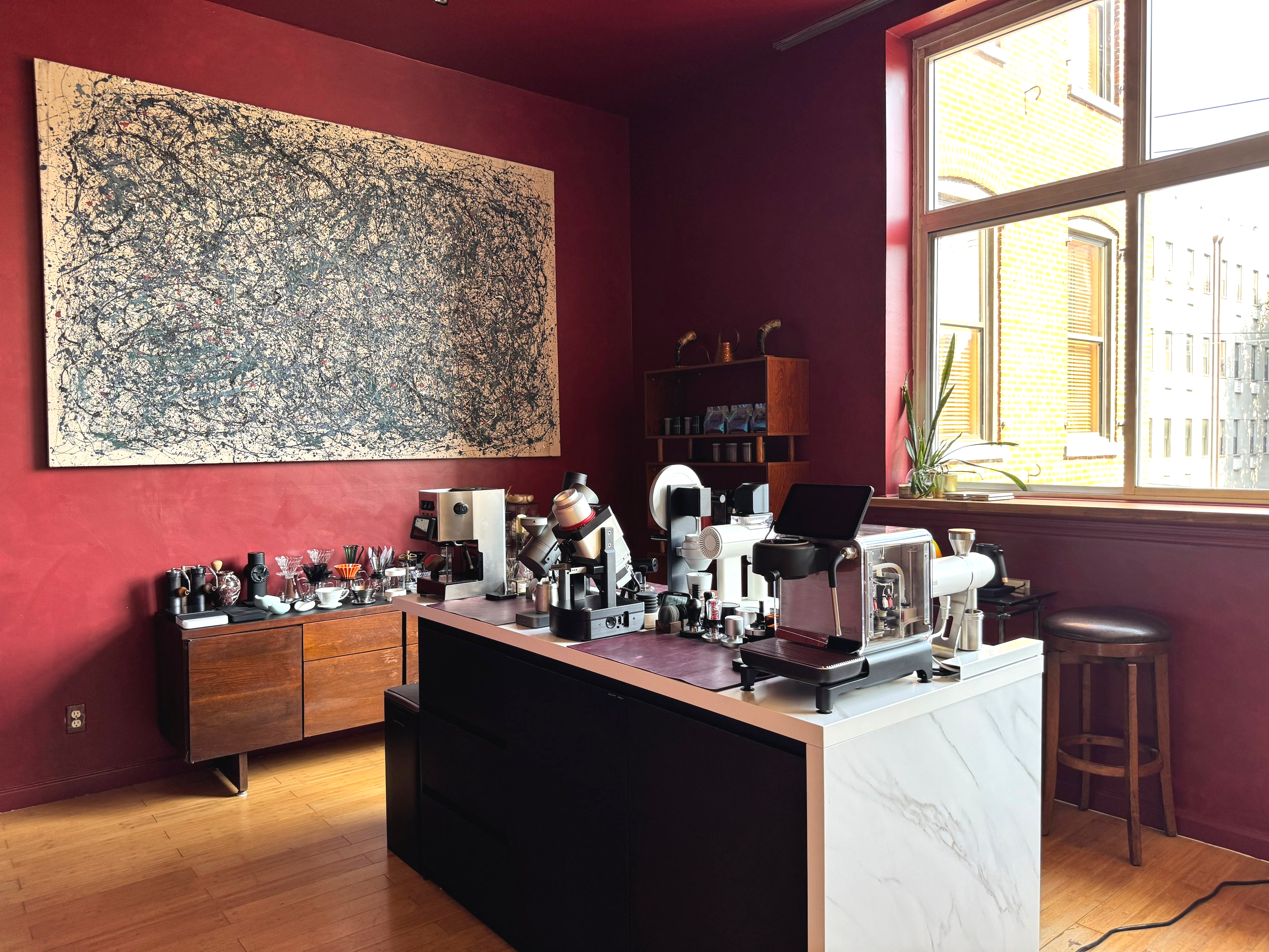 Coffee brewing equipment on a modern kitchen counter with a large abstract painting on a red wall and a window showing neighboring buildings.