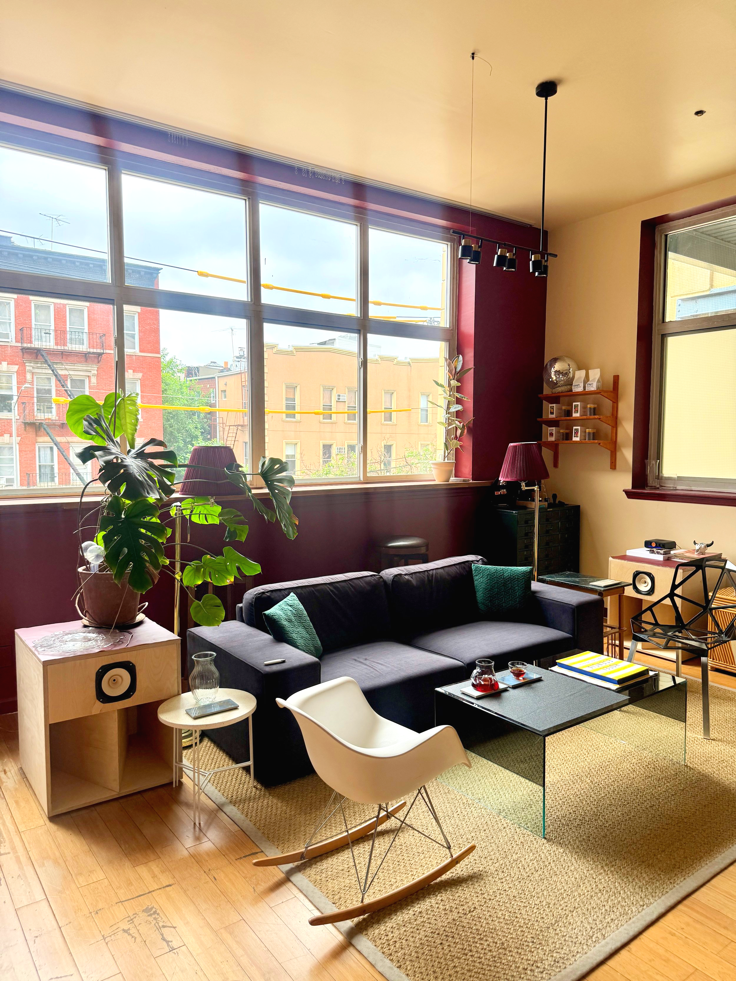 Living room with large windows, black sofa with green pillows, white rocking chair, glass coffee table with drinks, potted plant, side table, lamps, and decorative items.