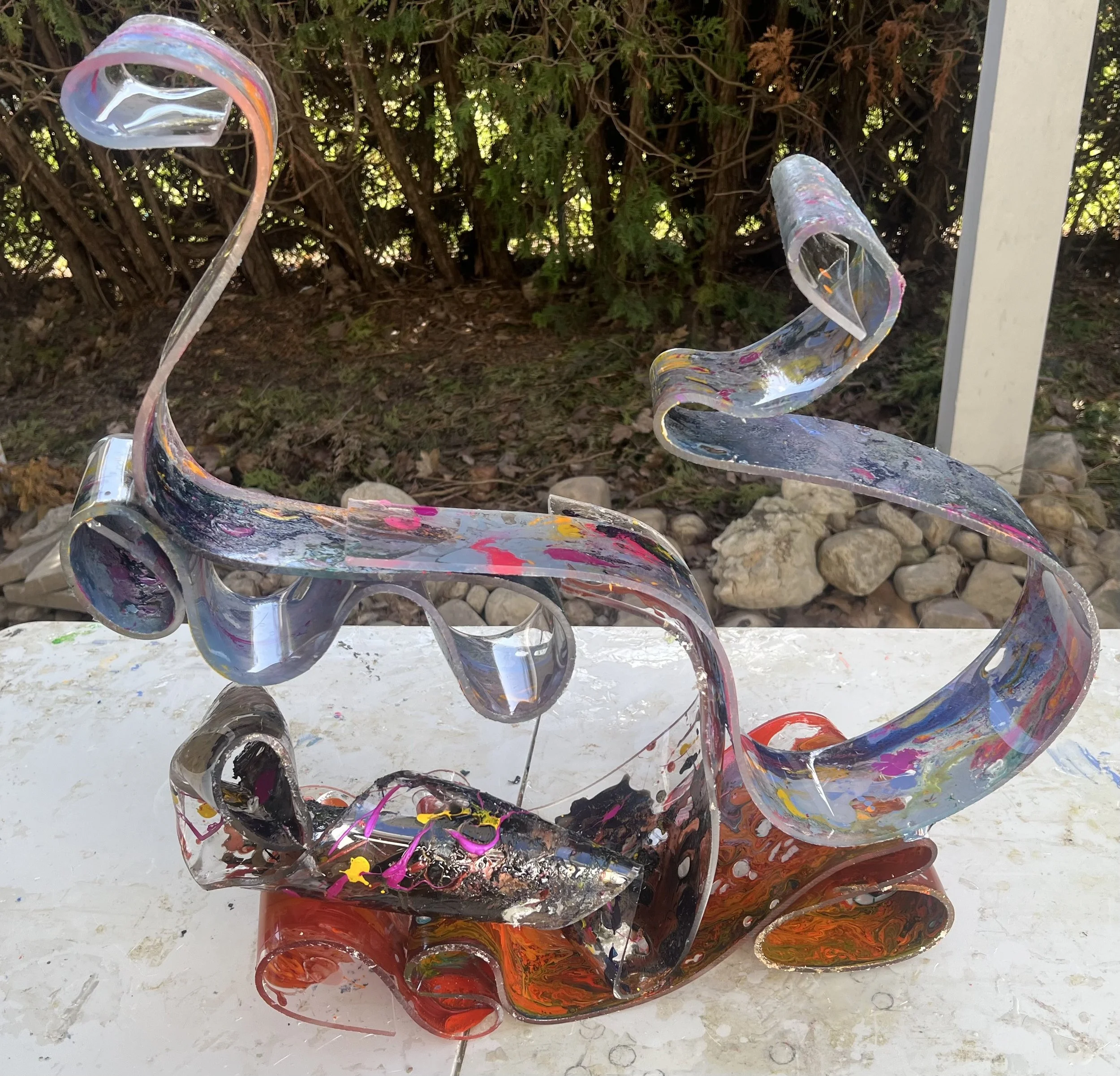 Colorful abstract sculpture with swirling, intertwined shapes, set outdoors with a background of dense green shrubbery and trees.