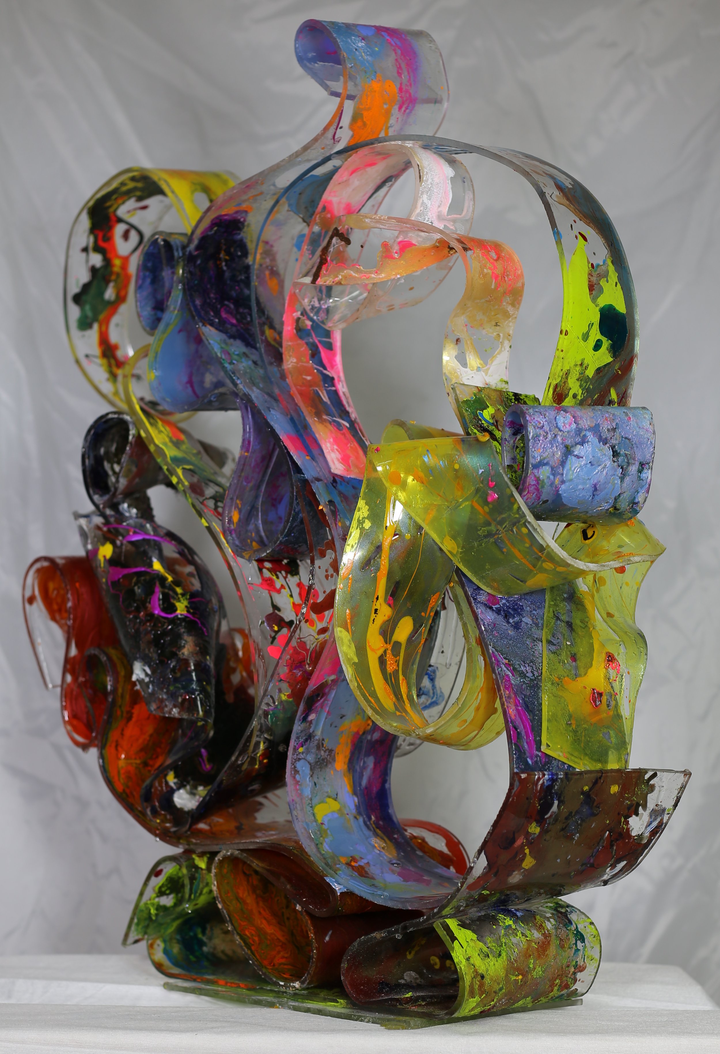 A colorful abstract sculpture made of interwoven, painted acrylic sheets with splashes of bright colors on a white surface with a plain background.