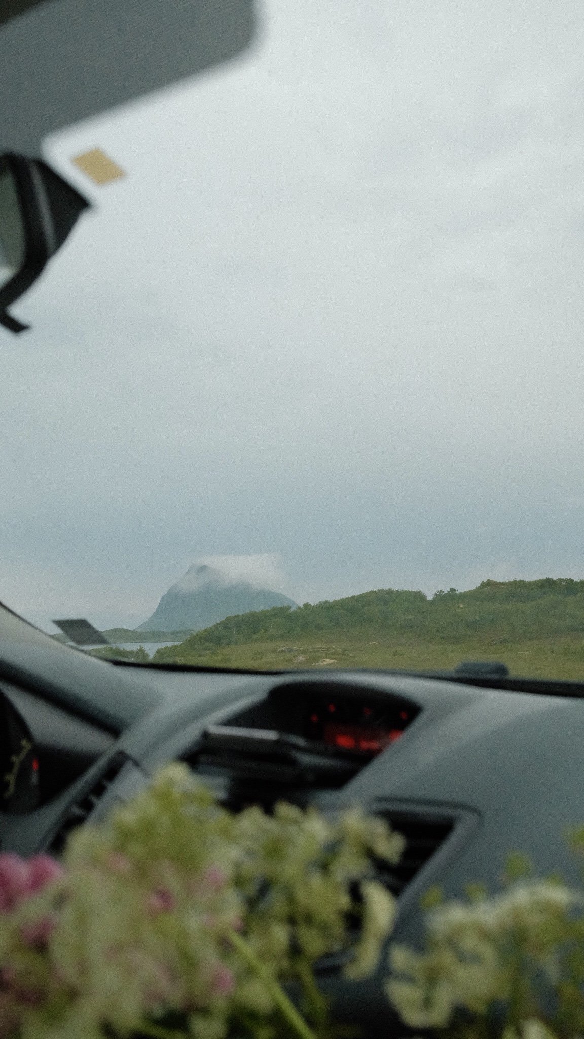 Utsikt fra bilen med fjell, vann og grønne landskap under en overskyet himmel.