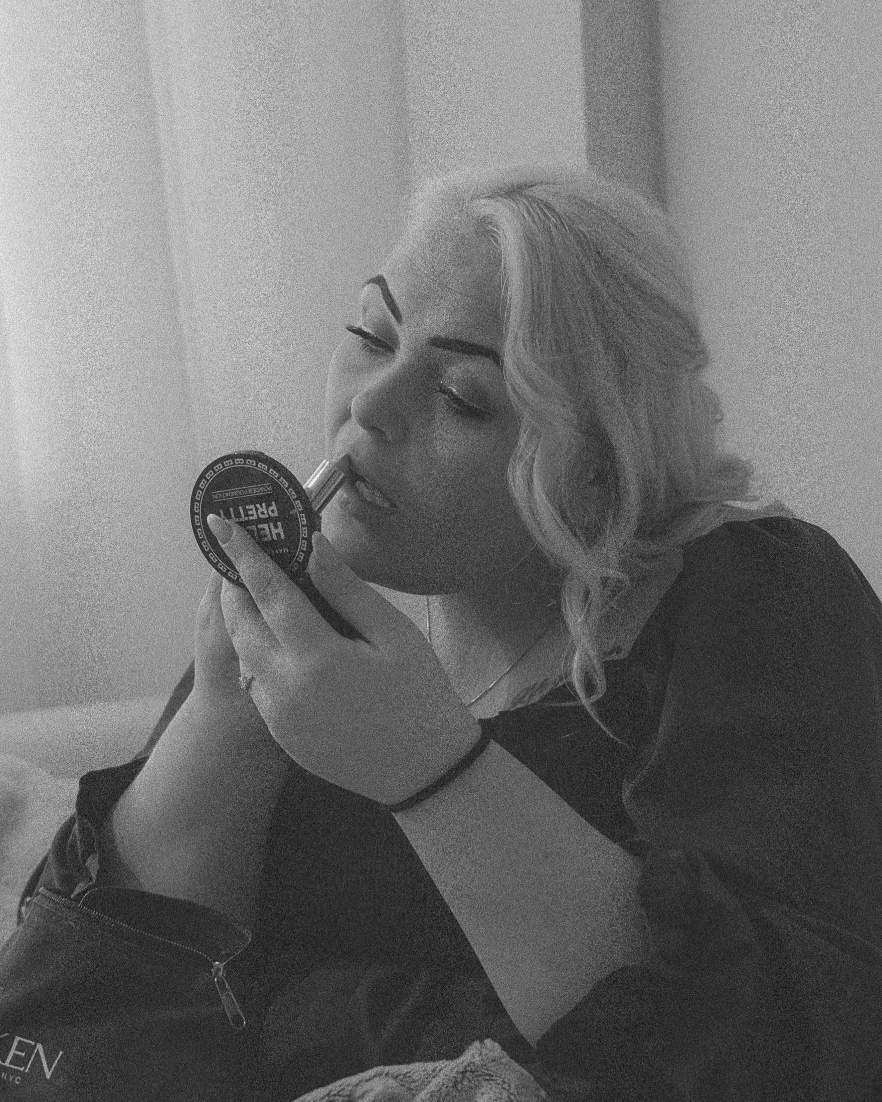 En kvinne med lyst hår som sminker seg med øyenskygge og leppestift, sitter oppreist i et rom.