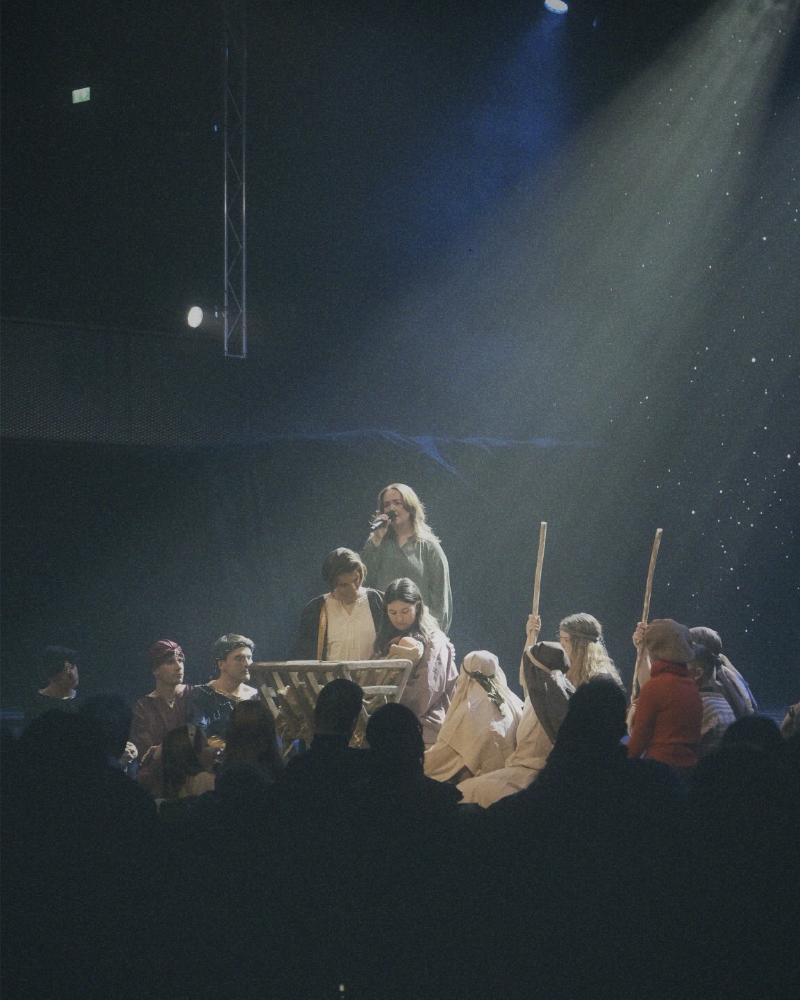 Et scenedrama med flere personer, der en kvinne står med mikrofon og ser ut til å snakke, omgitt av personer er kledd i tradisjonelle klær, noen med staver, og et publikum i forgrunnen.