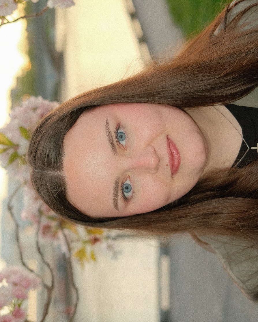 Nærbilde av en kvinne med langt brunt hår og blå øyne, tatt utendørs foran blomstrende trær.