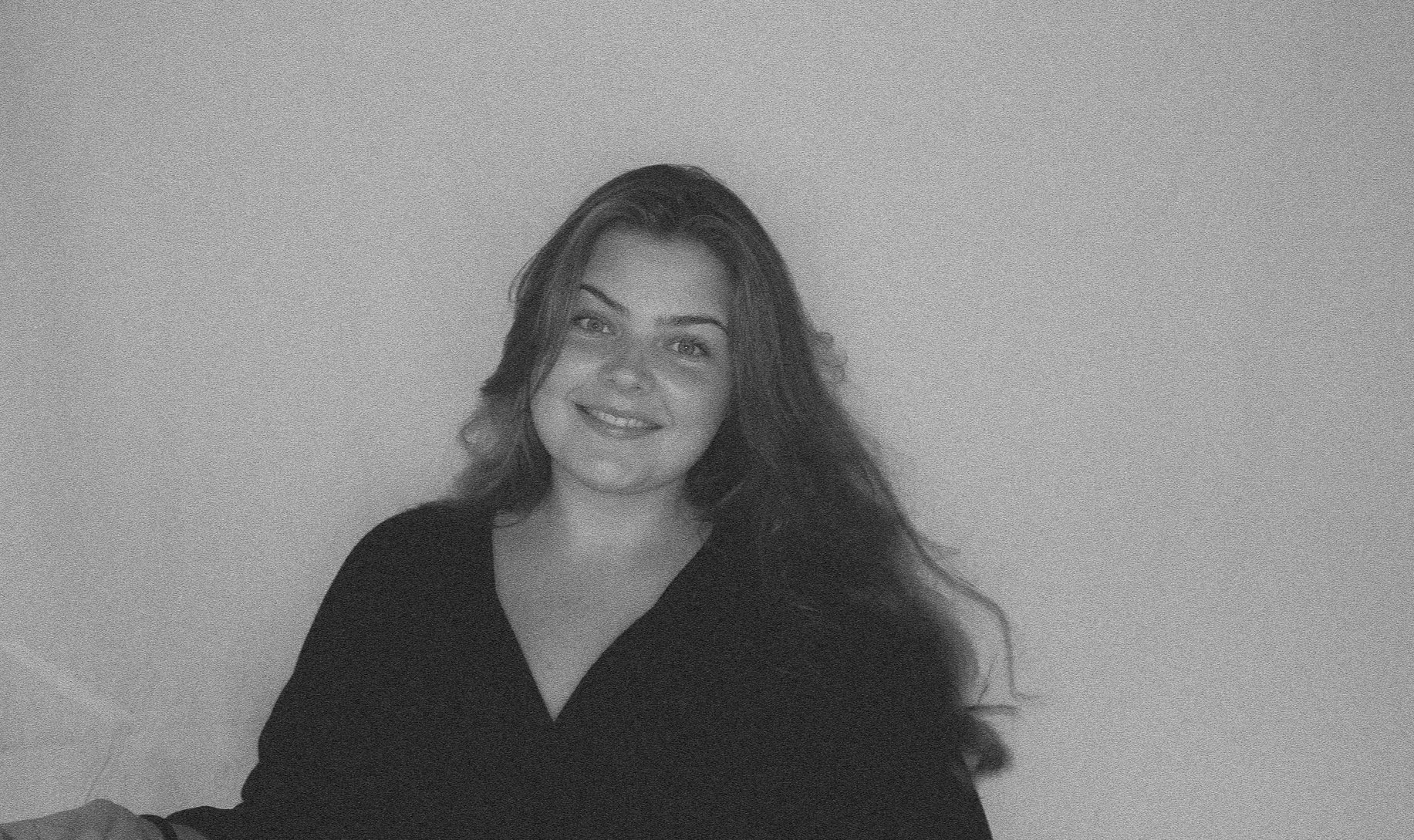 Et svart-hvitt bilde av en smilende kvinne med langt, bølget hår som har på seg en svart topp, opp mot en enkel, lys bakgrunn.