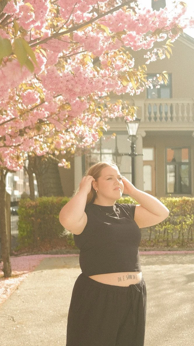 Kvinne i svart antrekk står under blomstrende kirsebærtre med rosa blomster, solskinn og en bygning i bakgrunnen.