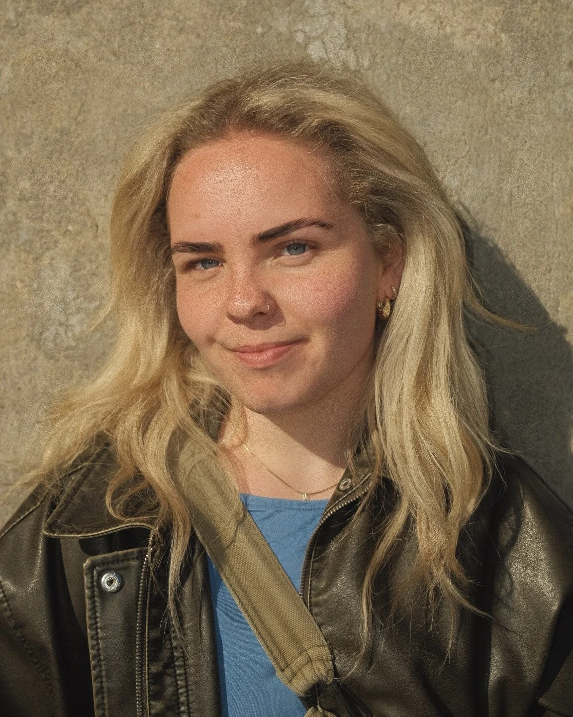 Et nærbilde av en ung kvinne med blondt hår, blå øyne, og lett smil, iført en skinnjakke og en blå topp, mot en nøytral vegg.