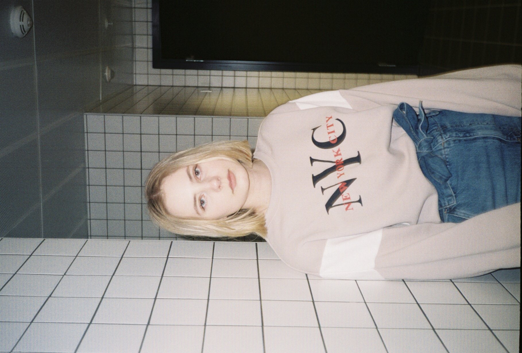 En ung voksen kvinne står mot en hvitkalket flisvegg, med en svart skjerm bak henne, iført en beige T-skjorte med 'New York City' skrevet på og blå jeans.