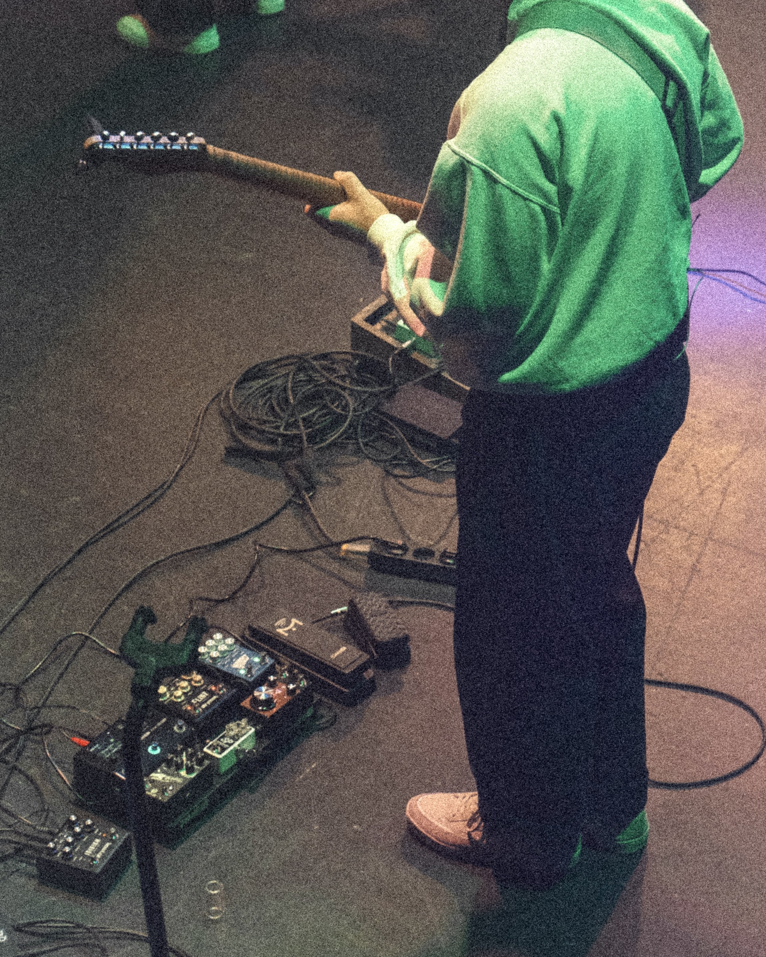 En person spiller på en elektrisk gitar med en pedalboard og andre musikkinstrumenter på gulvet.
