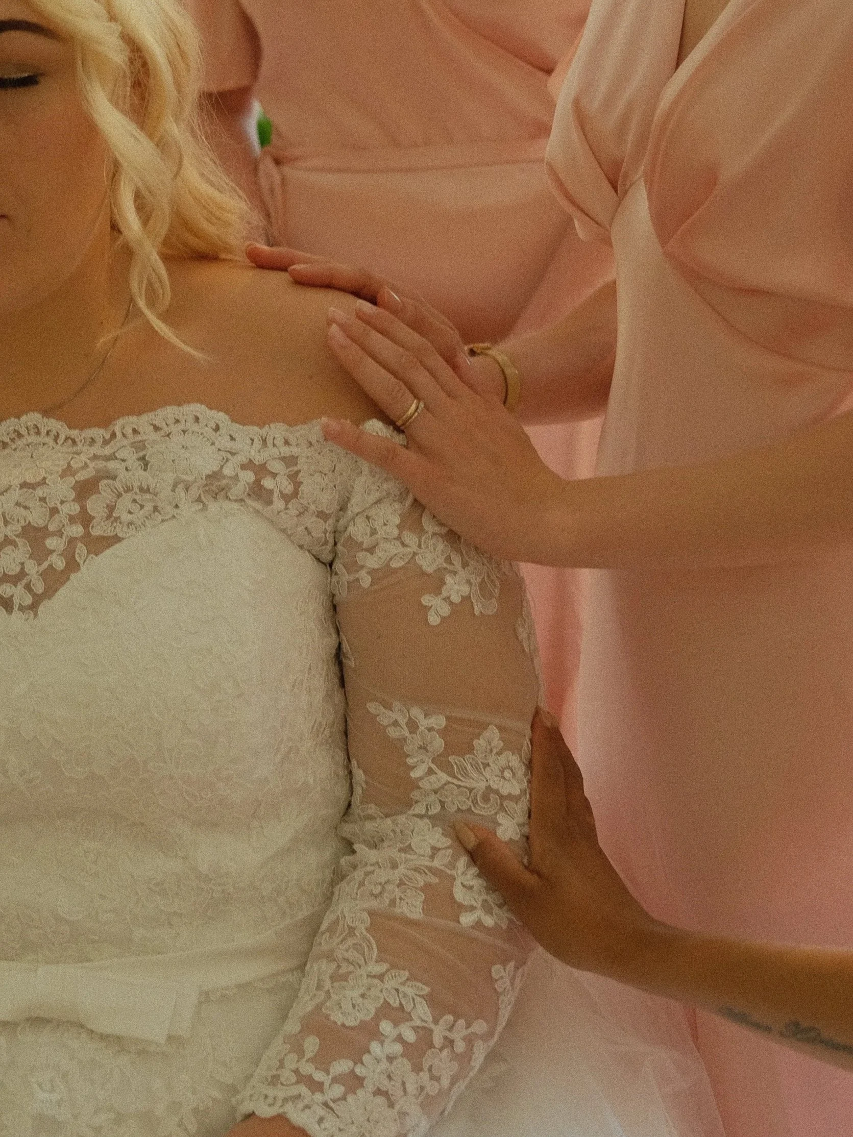 Nærbilde av en brud i hvit kjole med blonder, som får hjelp med å ta på seg kjolen av flere personer i rosa klær.