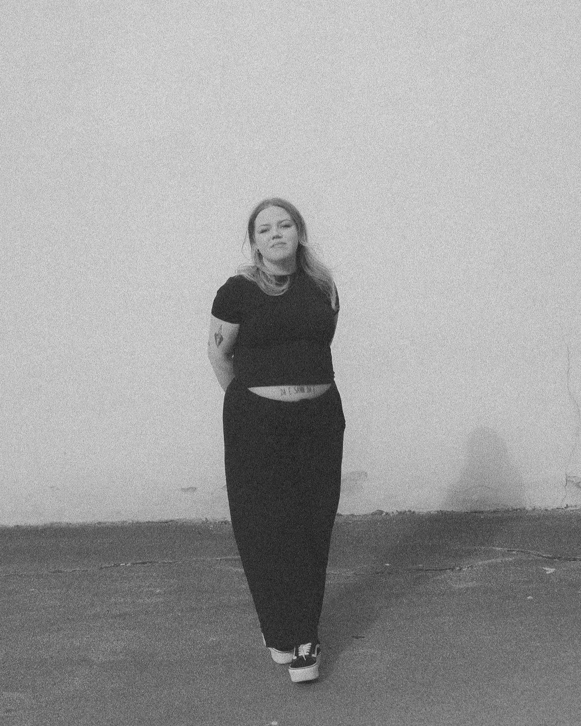 En kvinne står alene mot en tom, ulenkelig vegg, kledd i svart, med armlenker og sko med hvit såle.