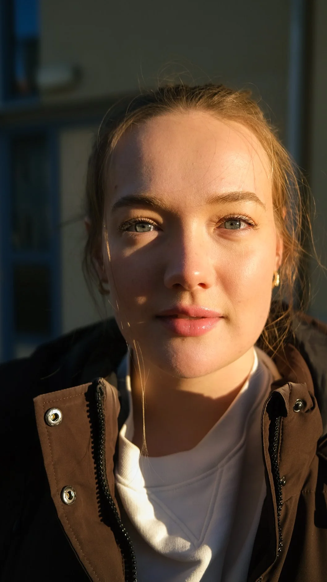 Nærbilde av en ung kvinne med lyse øyne og lyst hår, som ser mot kamera mens sollys belyser ansiktet hennes.