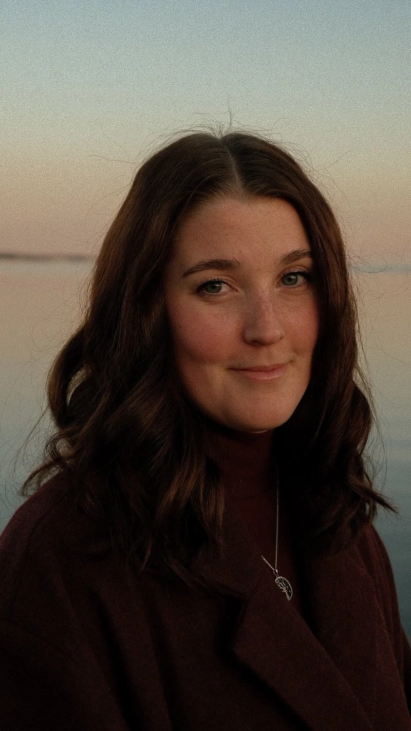 Portrett av en kvinne med langt brunt hår, iført burgunderfarget jakke og sølvhalskjede, mot en dus bakgrunn med en rolig sjø og himmel.