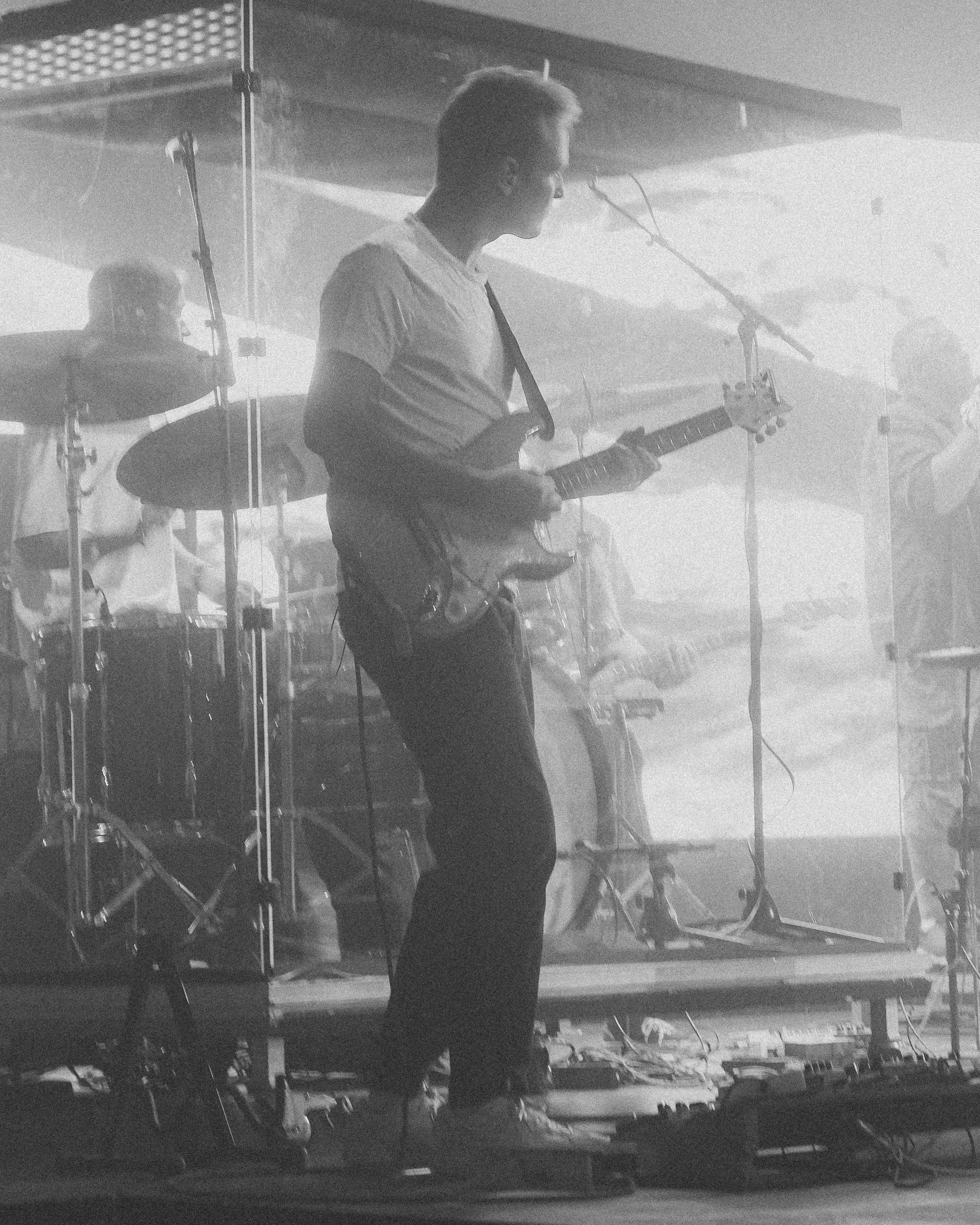 Musiker spiller elektrisk gitar på scene med trommeslagere i bakgrunnen, omgitt av musikkutstyr.