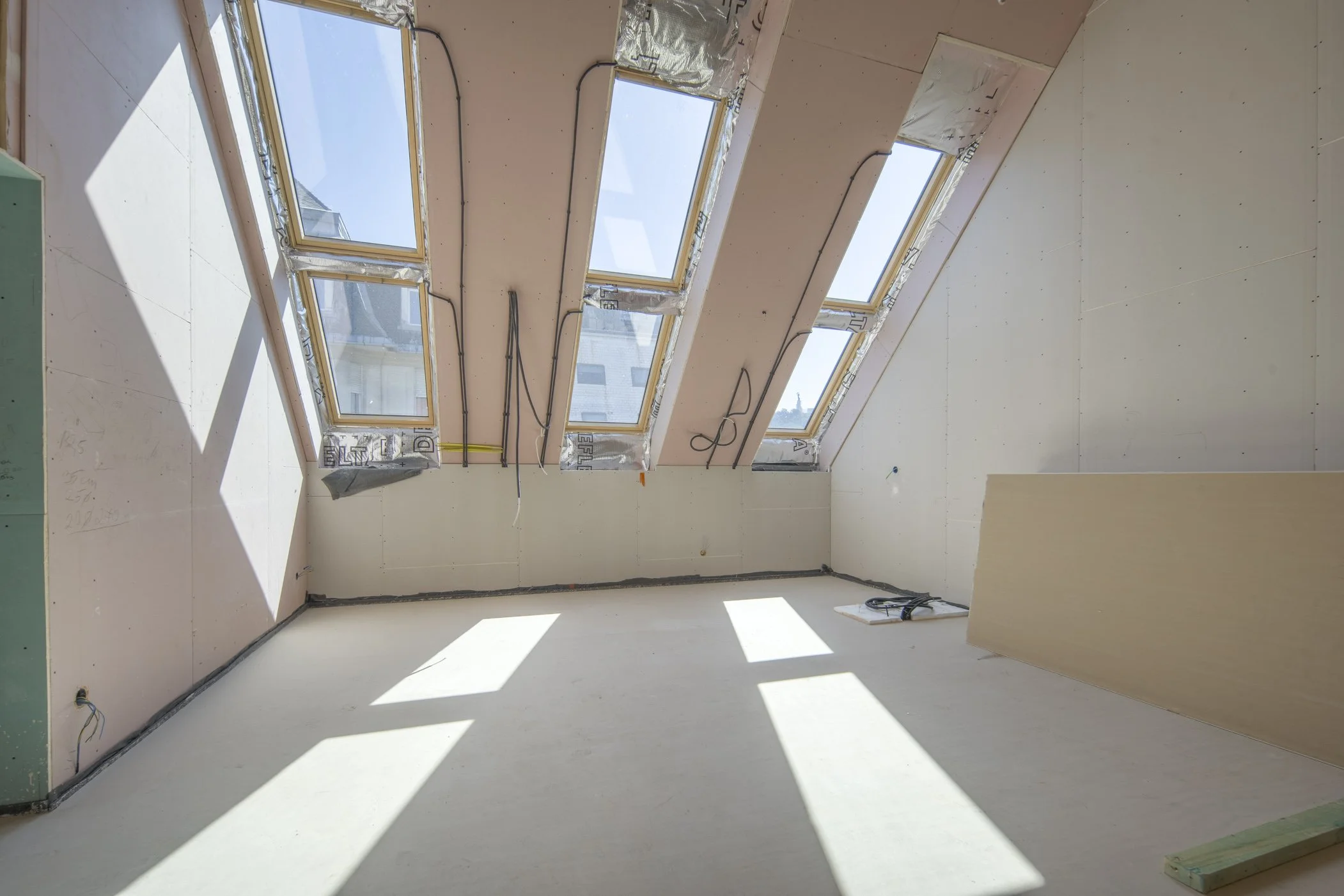 Építés alatt álló fényes helyiség, több nagy tetőtéri ablaknyílással és még építés alatt álló falakkal. A nagy üvegfelületeken besüt a nap és jól látszik a szemközti épület valamint a Gellért-hegy és a Szabadság-szobor is.