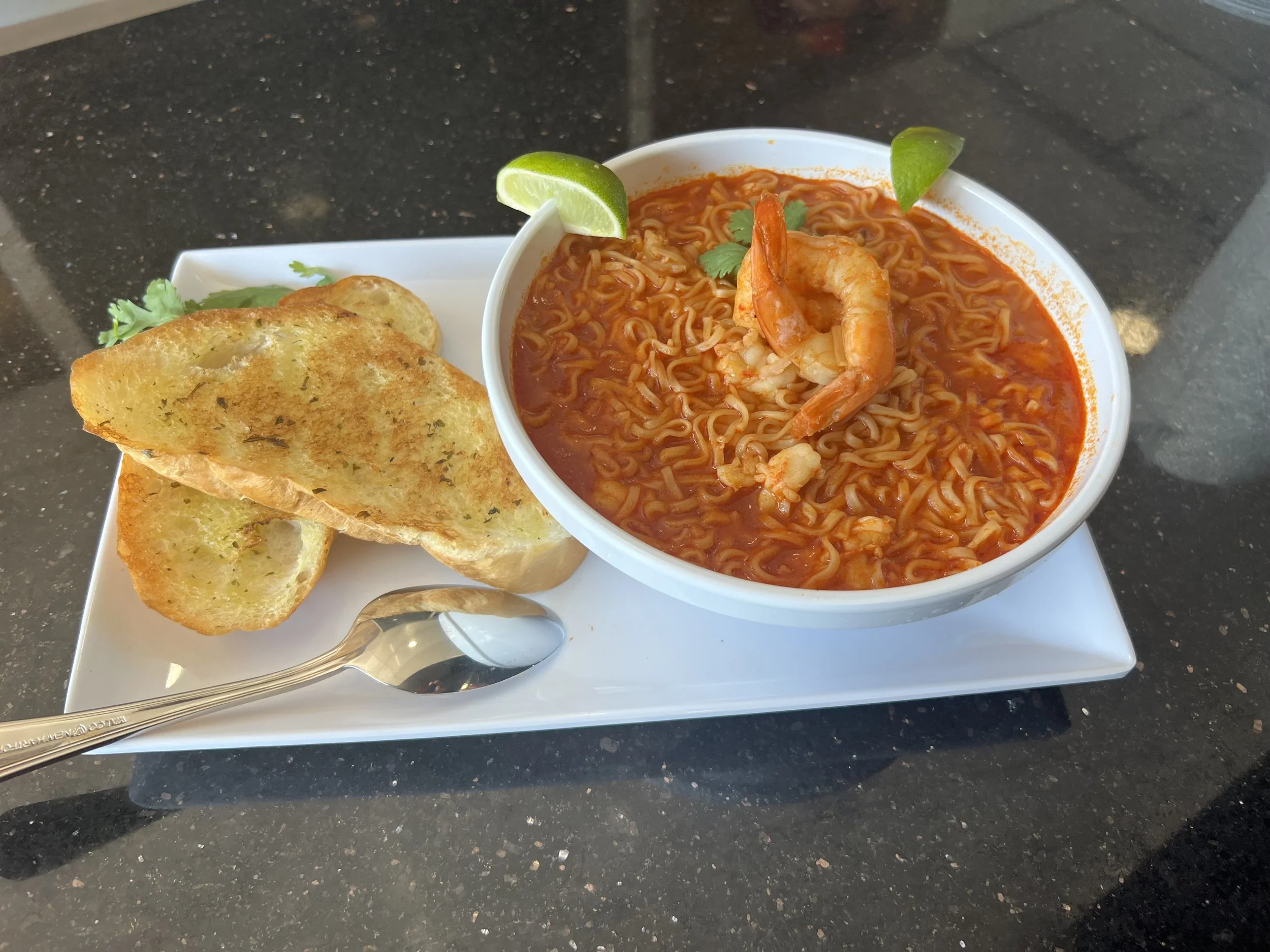 Bowl of spicy shrimp ramen with lime wedge and cilantro, served with garlic bread on a white rectangular plate.