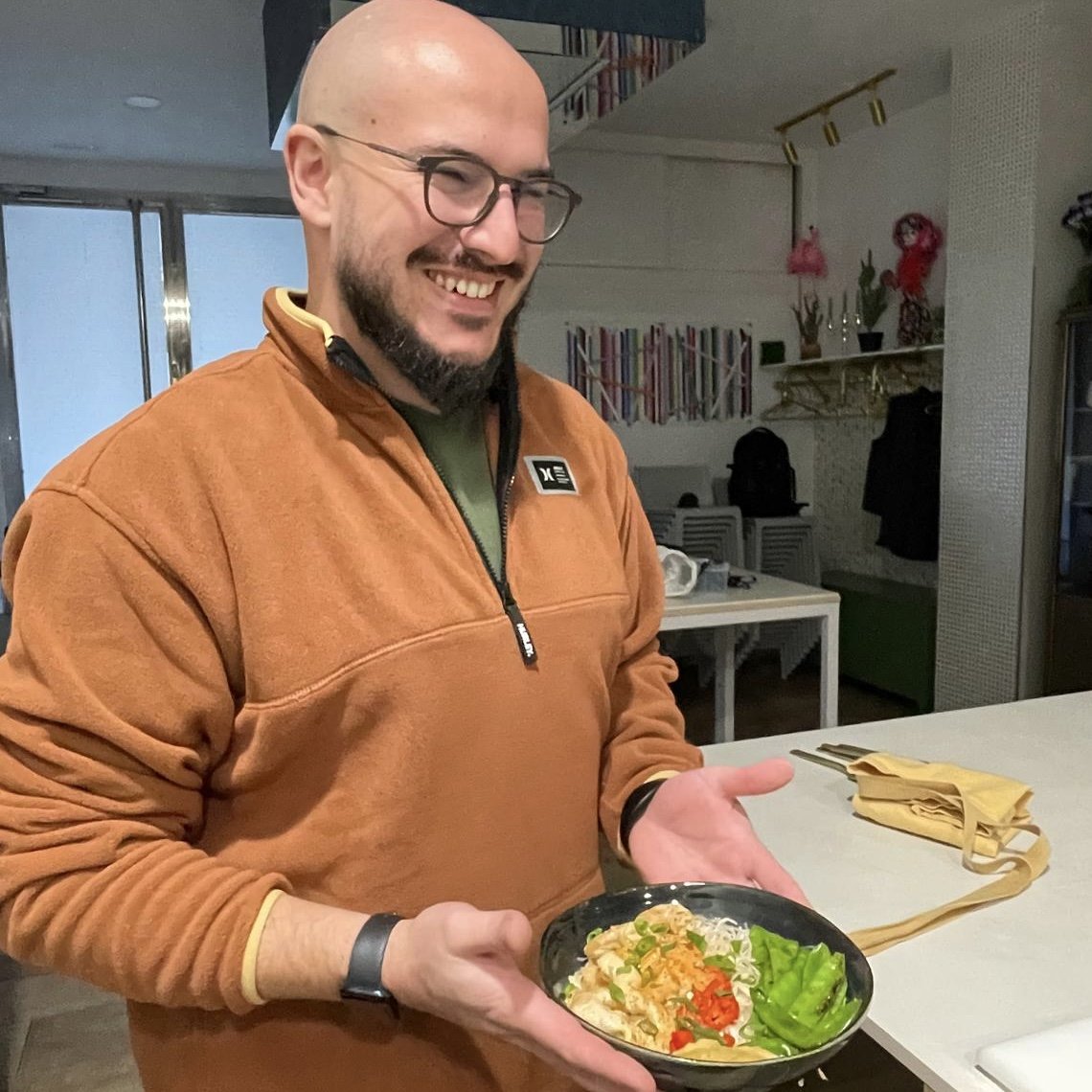 Smiling Carlso holding a bowl of food with rice, sliced red and green vegetables, and garnishes in a kitchen setting.