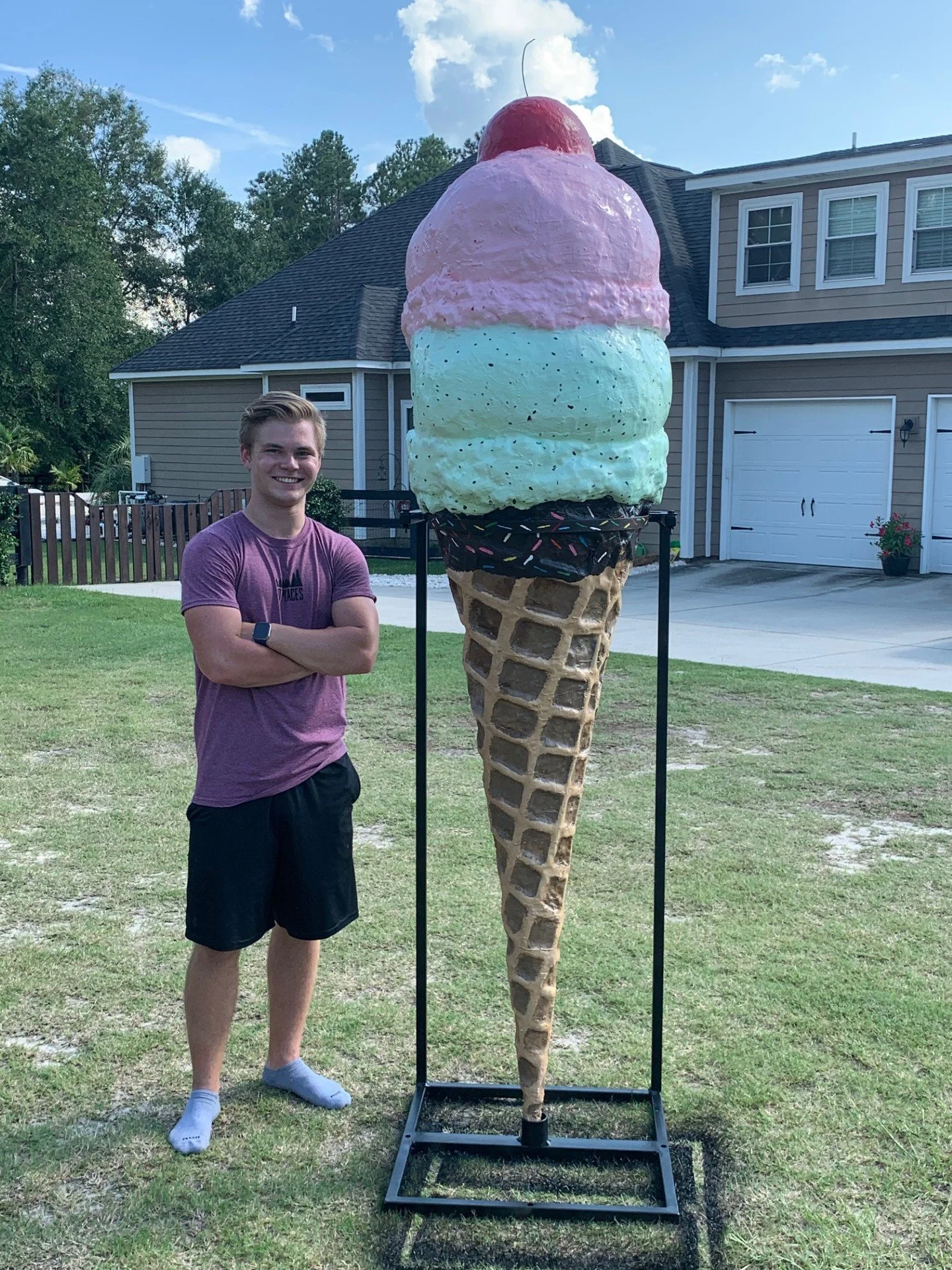 Man standing next to a large ice cream cone sculpture with pink and green scoops, a cherry on top, and sprinkles, on a grass lawn with a house in the background.