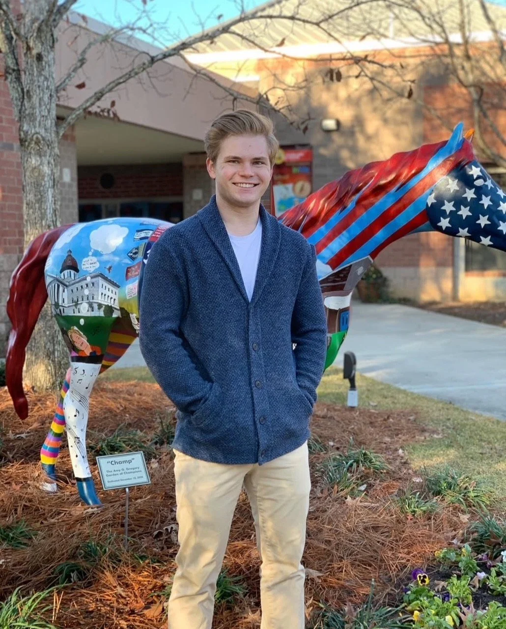 A person standing outdoors in front of a painted horse sculpture, wearing a blue sweater and beige pants. The background includes a building and trees.