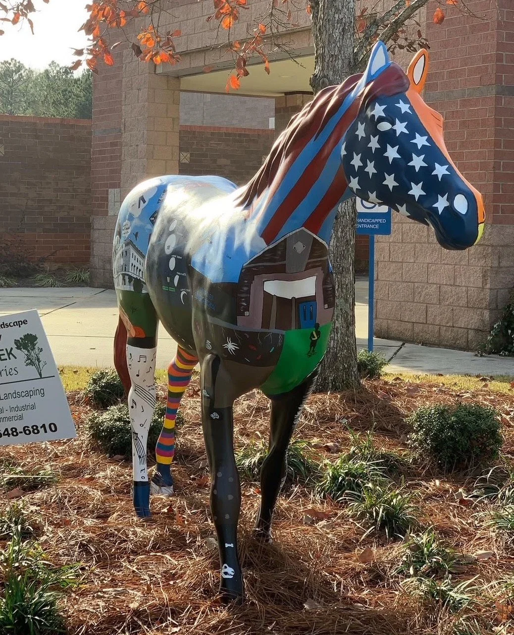 Colorfully painted horse sculpture with an American flag pattern on head and diverse illustrations on the body, displayed outdoors.