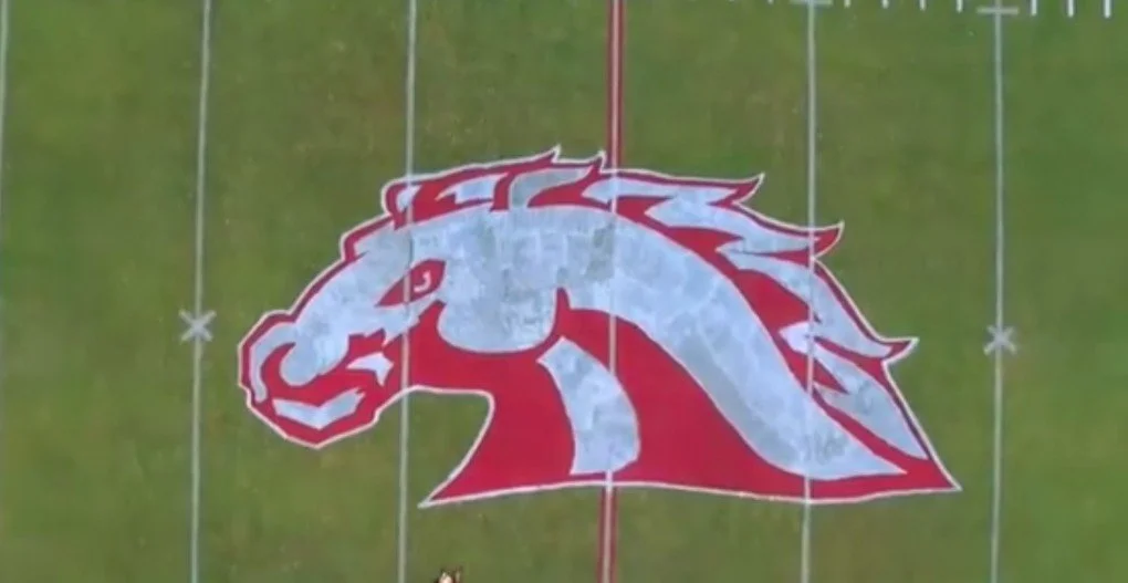 Aerial view of a football field with a large mascot logo painted at the center, depicting a stylized horse head in red and gray.
