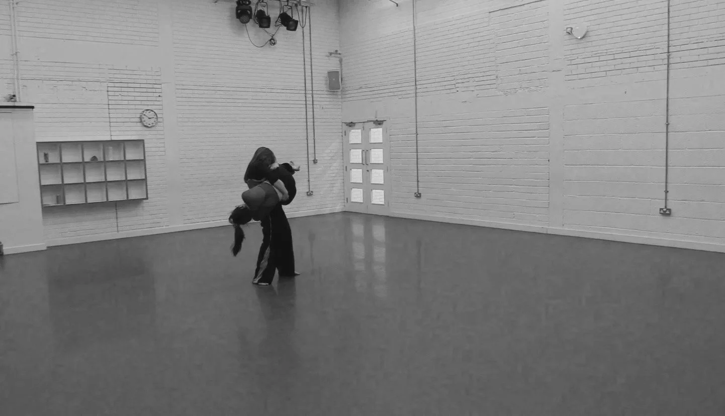 In a studio, two girls perform a koala style lift in which one holds the other up around the waist and legs whilst the other lays across them wrapped around their torso