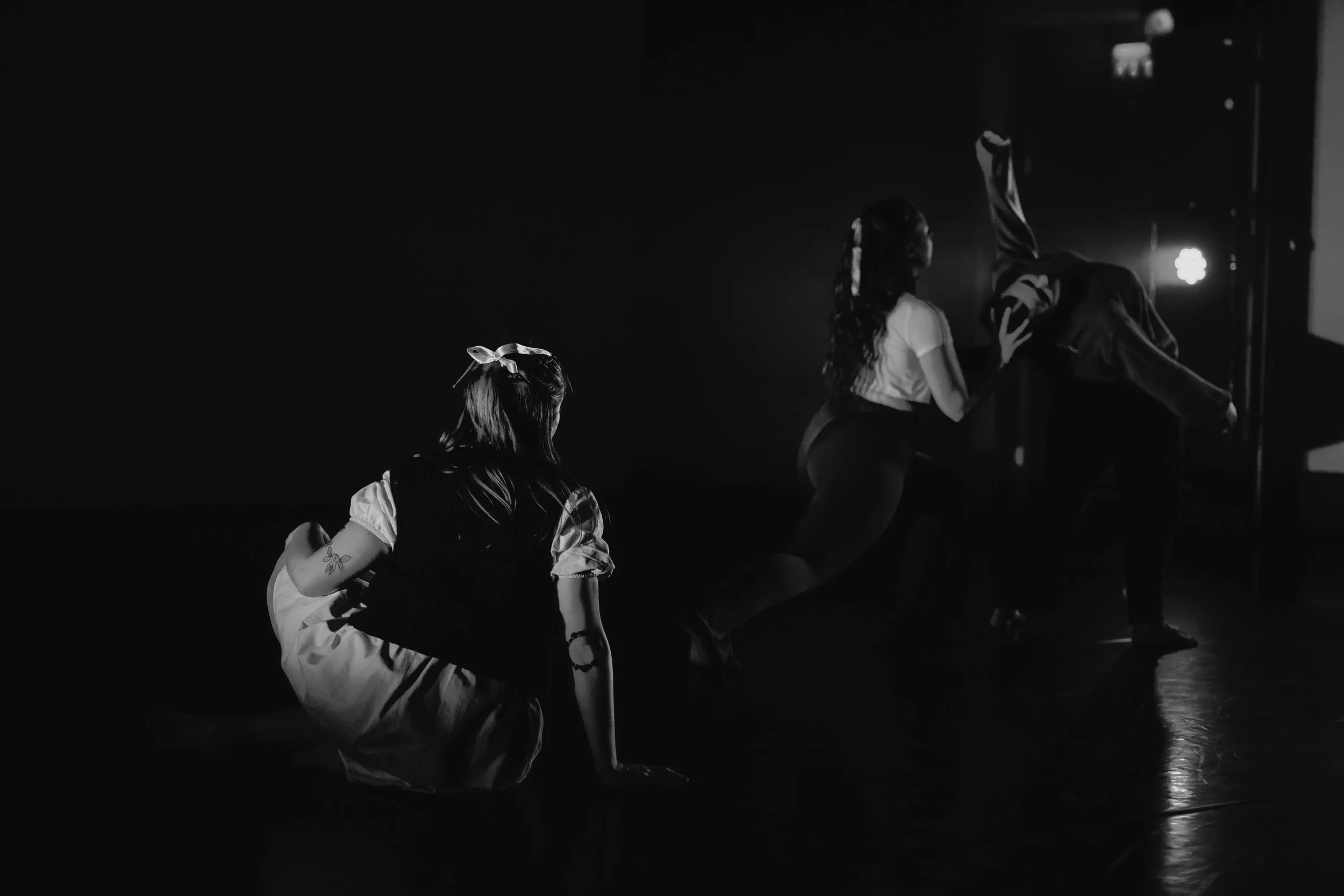In black and white, one dancer is sat on the floor in a white dress and knitted vest, looking over at a duet of dancers. In the duet, one dancer arches back whilst the other lunges and holds and assists the other's head and back.