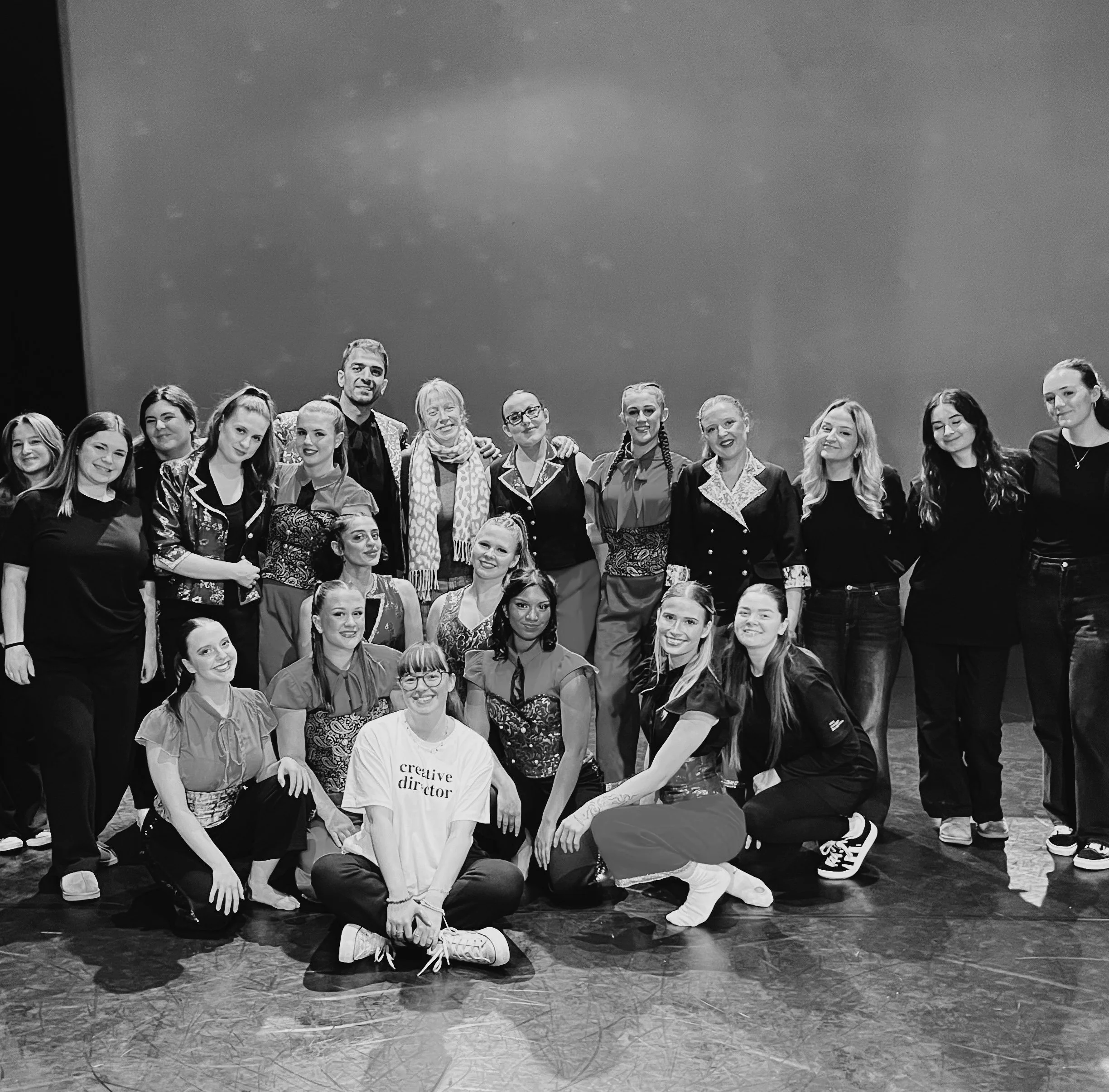 A group of dancers and technicians pose together, some sat, some stood in a clump. They all smile to the camera.