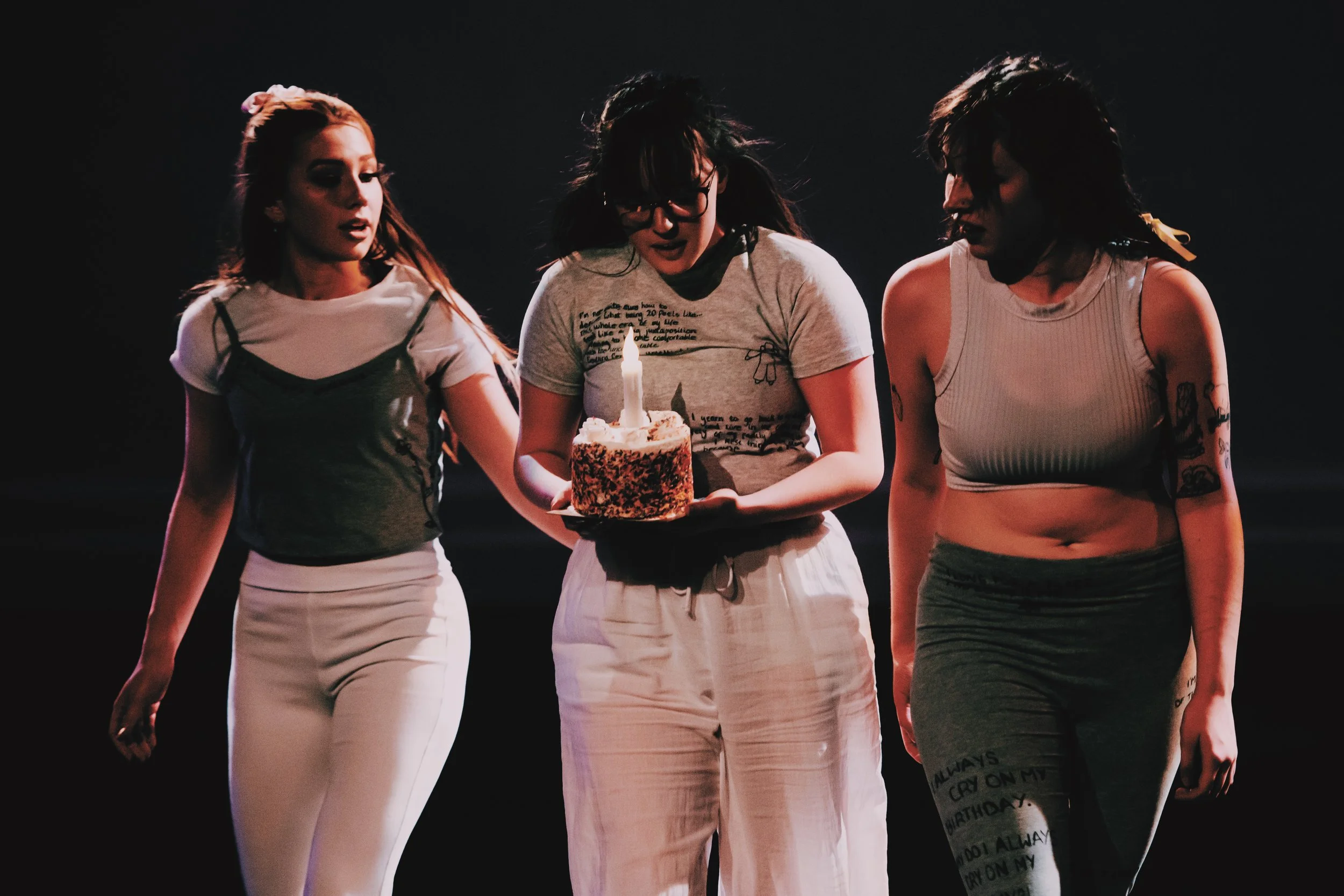 Three dancers are walking forwards in a shadowy stage. They are wearing grey and white clothes and their hair flows with the motion of them moving. The central girl is holding a birthday cake with a rainbow sprinkle coating and a big lit candle.