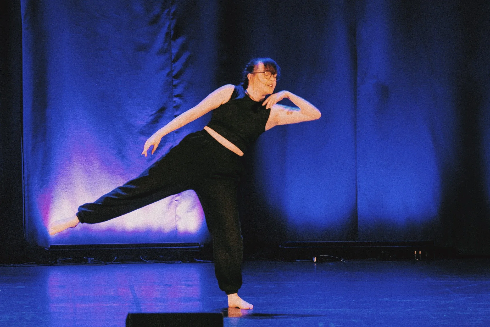 On a stage backlit by blue lights, and a warm front light, a dancer in all black, hair plaited, is mid motion, circling their right elbow and shoulder, leaning off to that side, with the opposing leg extended out with the feet pointed