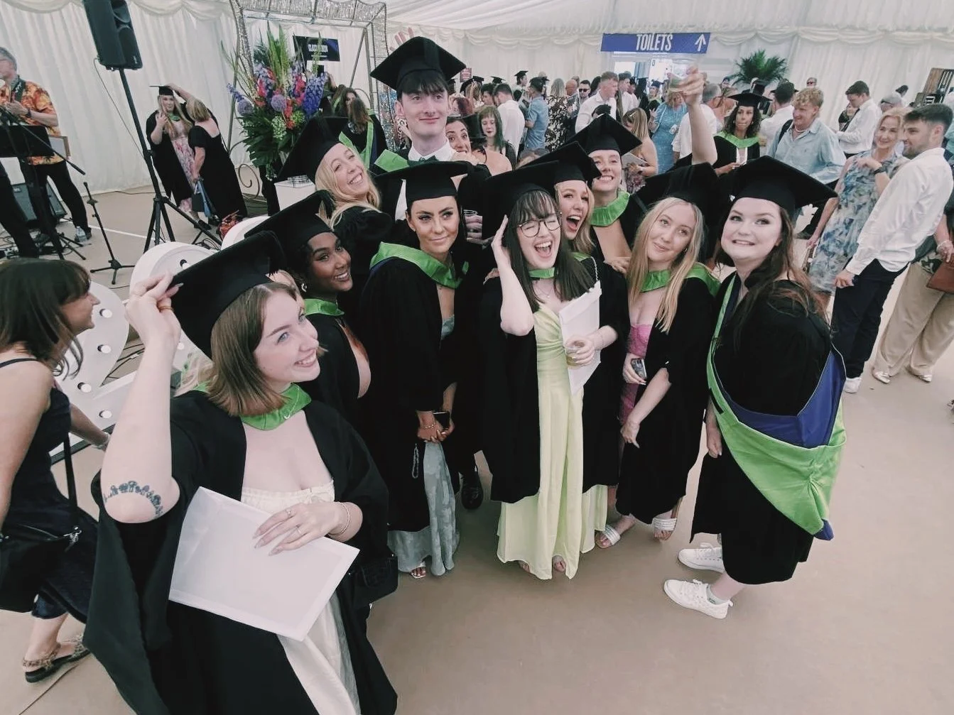 A group of people pose in celebration at graduation. They are huddled together in caps and gowns and all look very joyous.