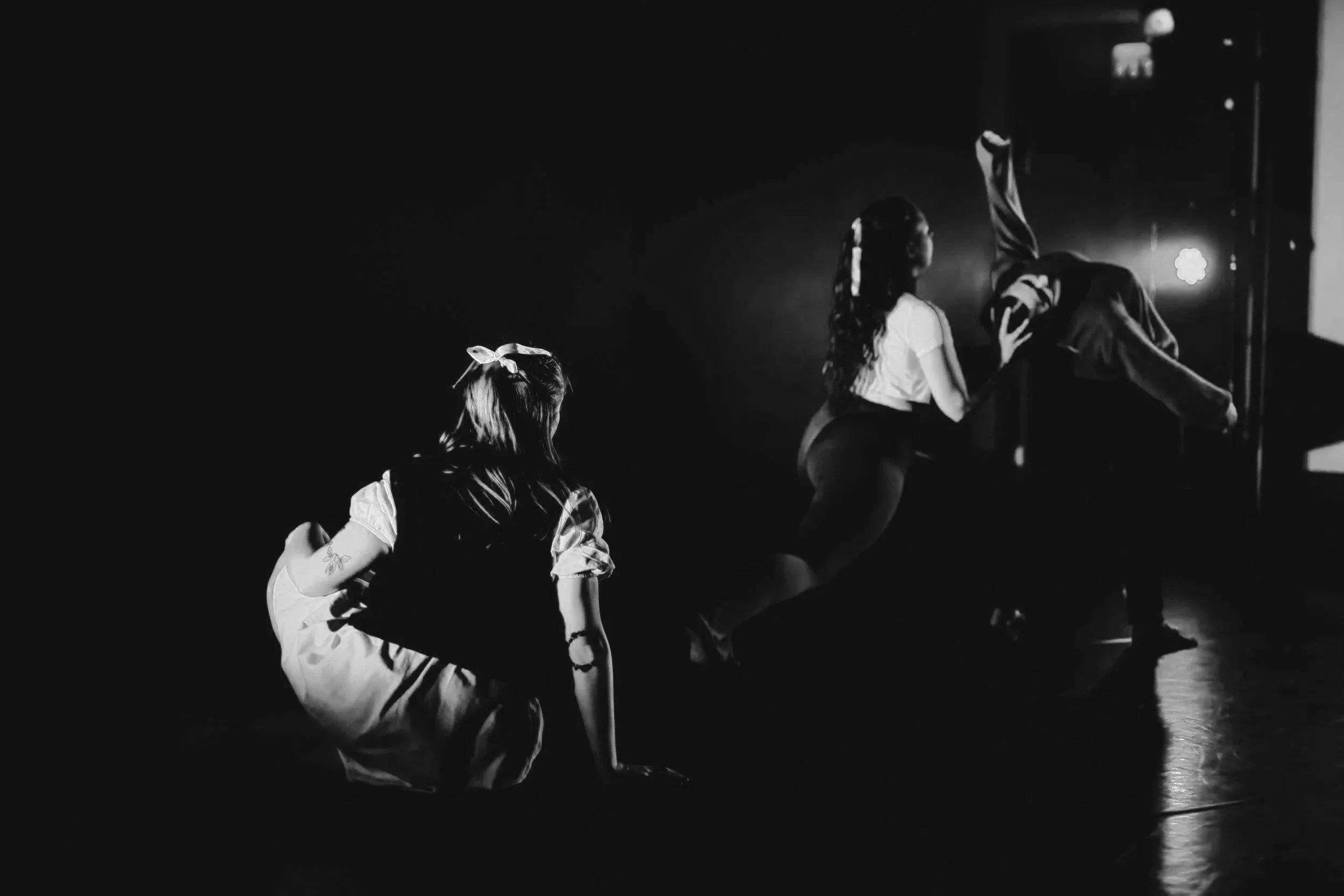 In black and white, one dancer is sat on the floor in a white dress and knitted vest, looking over at a duet of dancers. In the duet, one dancer arches back whilst the other lunges and holds and assists the other's head and back.