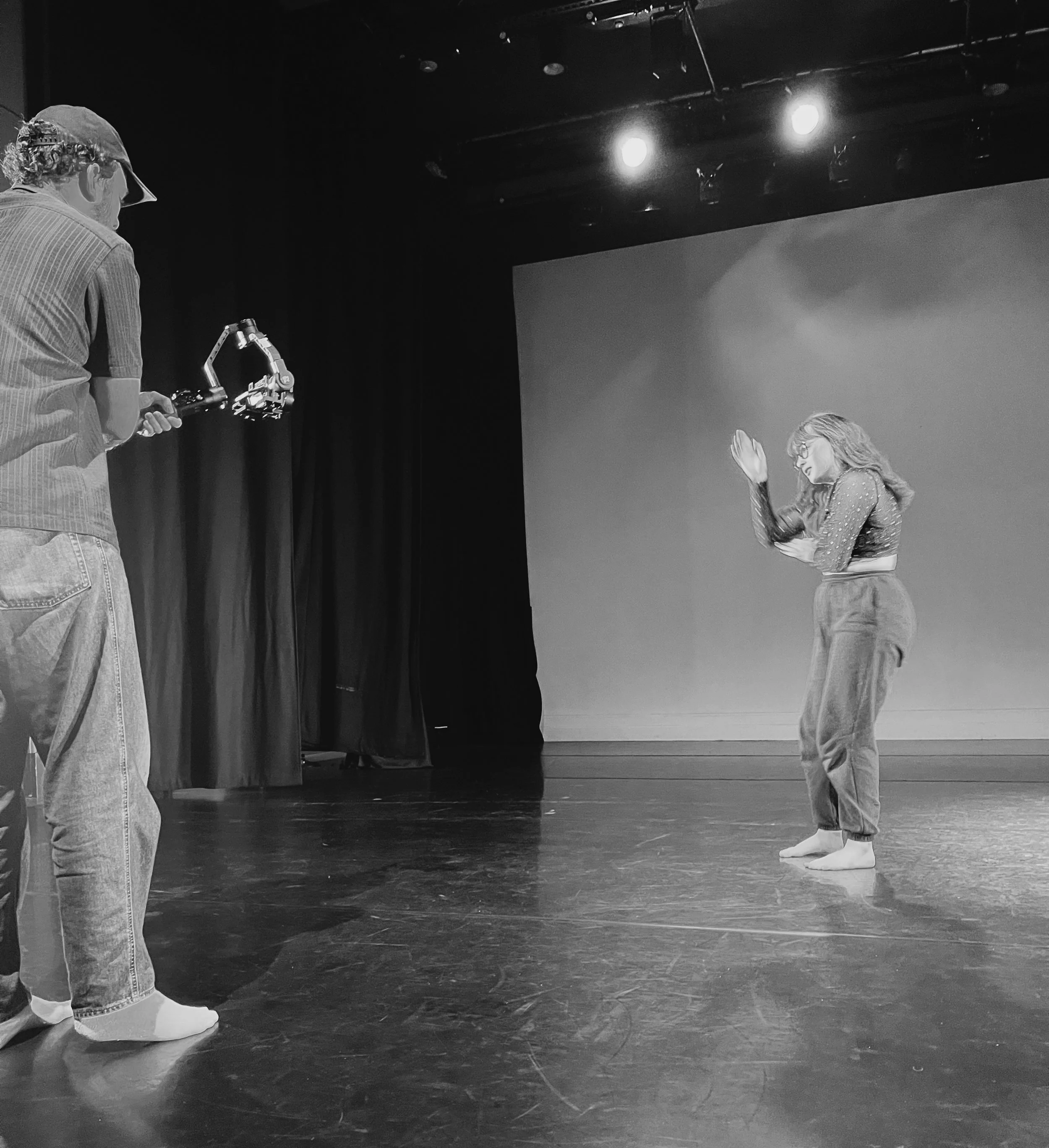 A camera man with a mobile rig stands close to the left side of the image filming the dancer lit by an orange glow. The dancer 's right arm is palm up near her face pulling downwards which will have a knock on effect to move her body backwards.