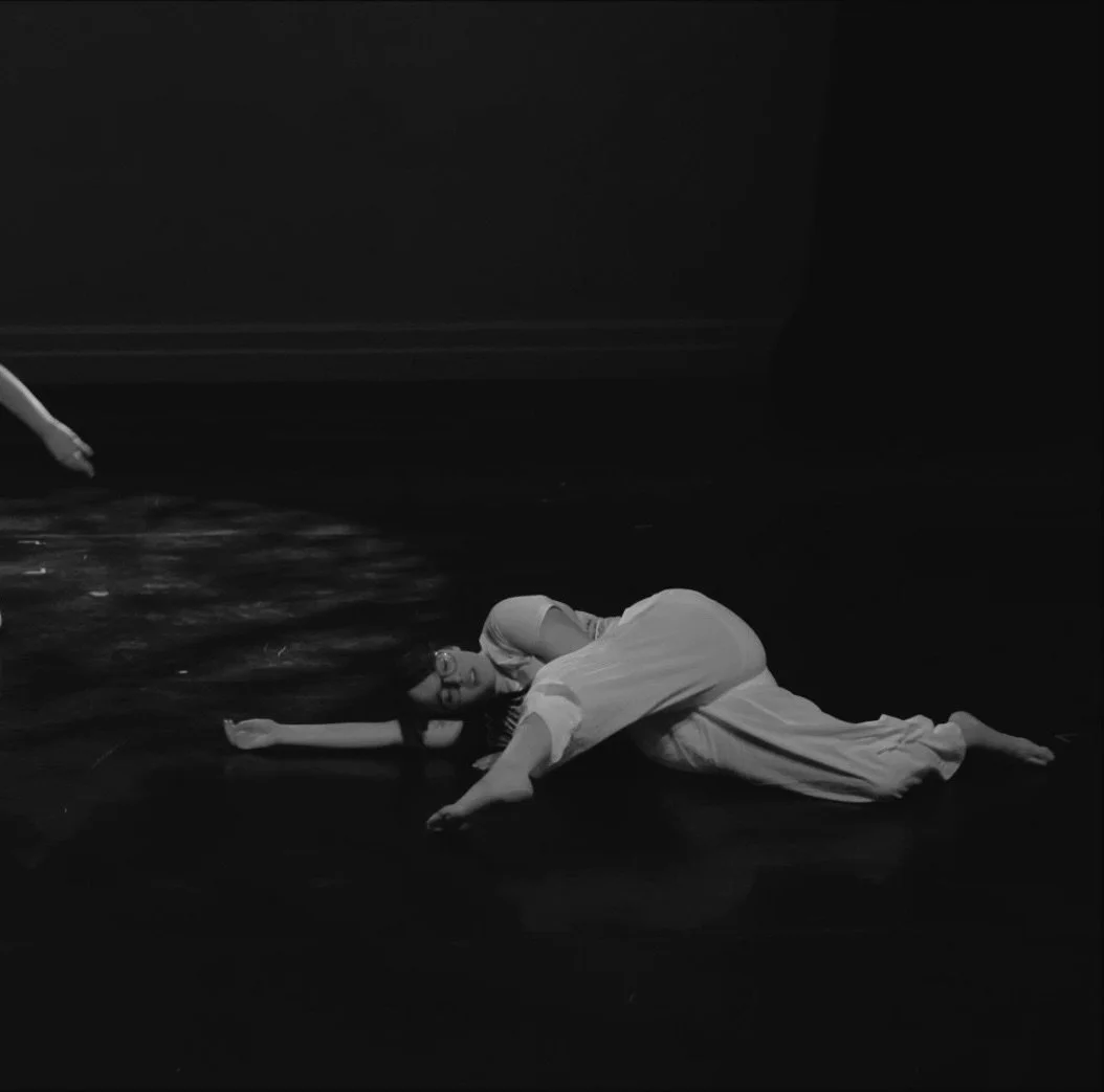 Lying on the floor of the stage, a dancer has her arm outstretched with her head laying ontop of it on her side as her leg swings forward in front of her.