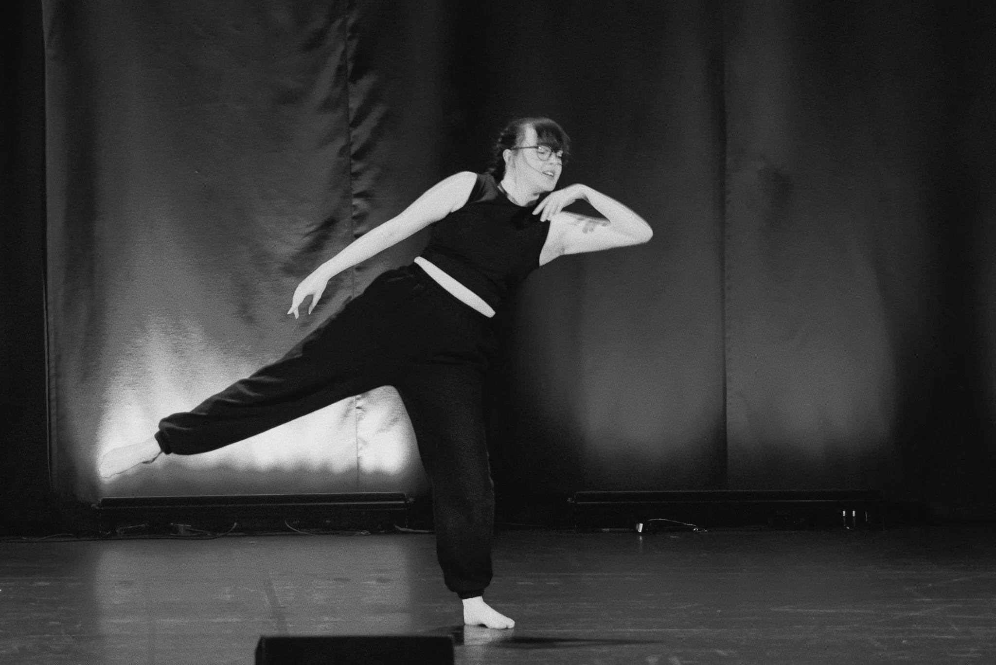 A camera man with a mobile rig stands close to the left side of the image filming the dancer lit by an orange glow. The dancer 's right arm is palm up near her face pulling downwards which will have a knock on effect to move her body backwards.