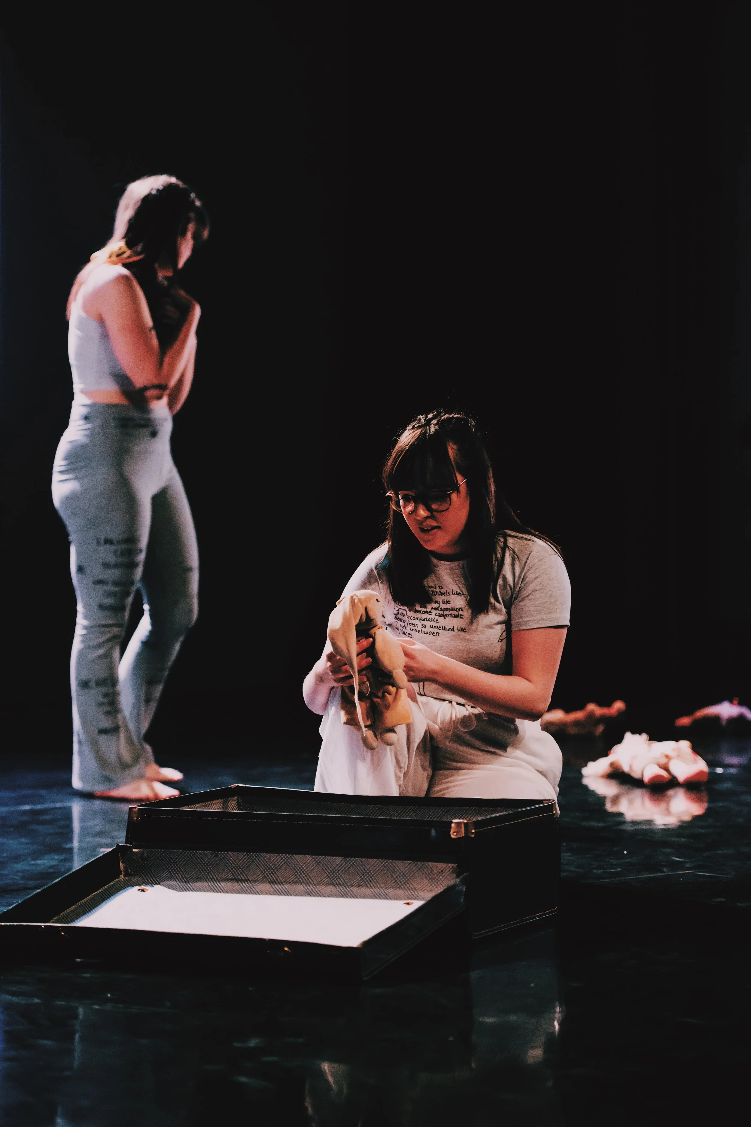 Two dancers perform on a dim stage. One sits on the floor unpacking a soft teddy from an open suitcase, while the other stands in the background, looking away with contemplative posture. Scattered plush toys lie nearby.